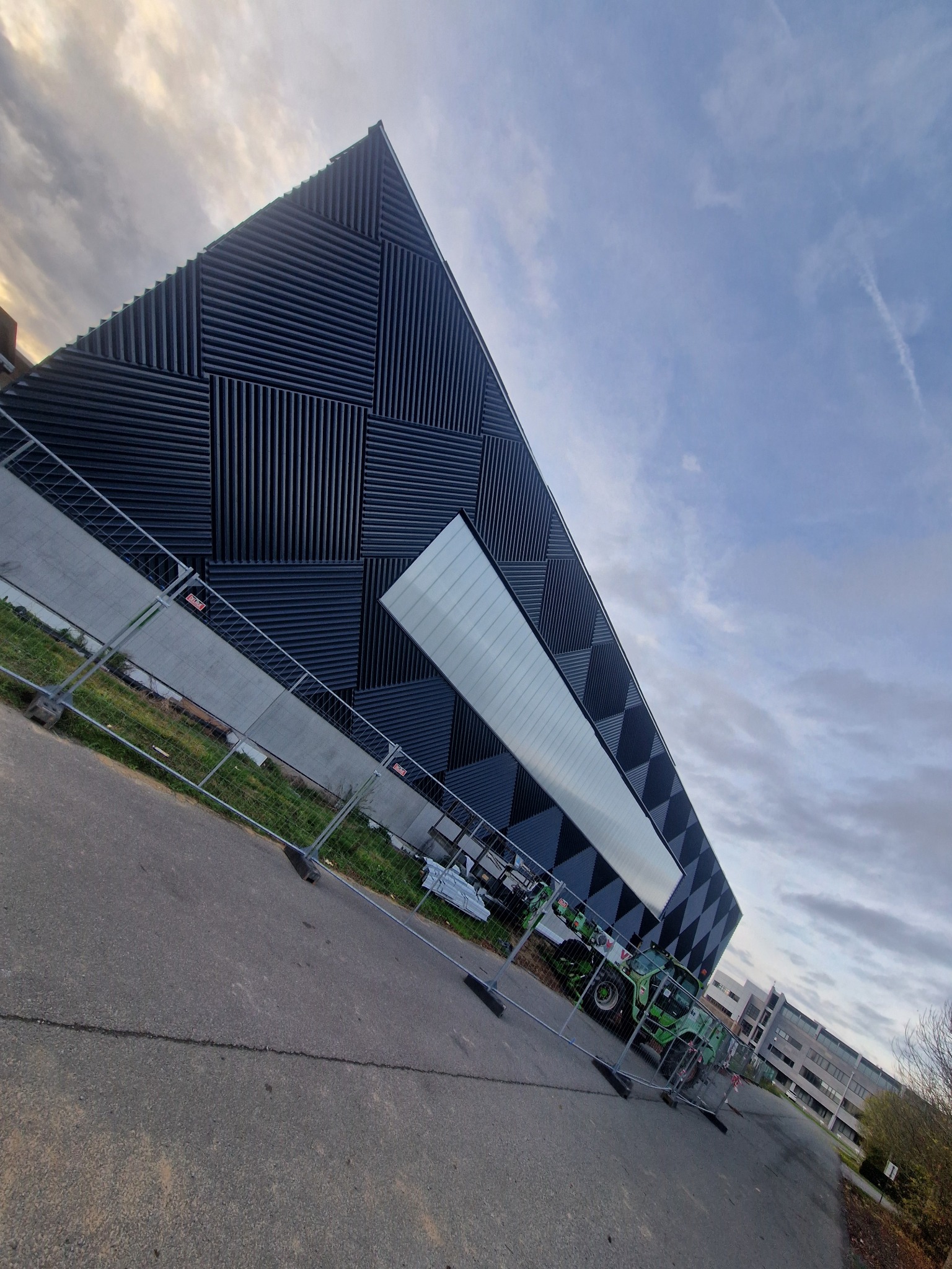 Nowoczesna hala z czarną, wzorzystą elewacją z płyt warstwowych, widziana z dołu pod kątem. Na pierwszym planie barierki i maszyna budowlana.