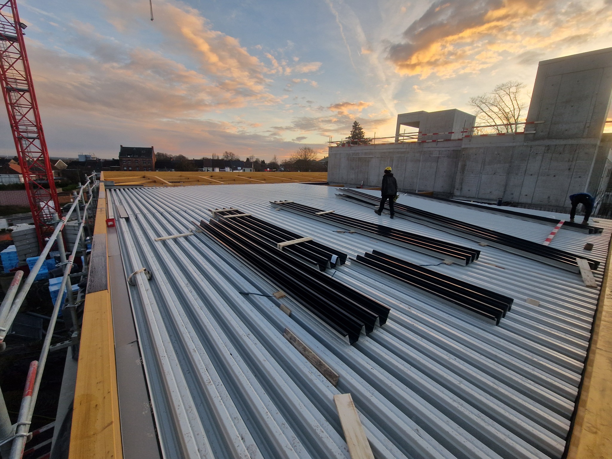 Montaż blachy trapezowej na dachu hali w trakcie budowy. Widoczni pracownicy w kaskach i elementy konstrukcyjne na tle zachodzącego słońca.