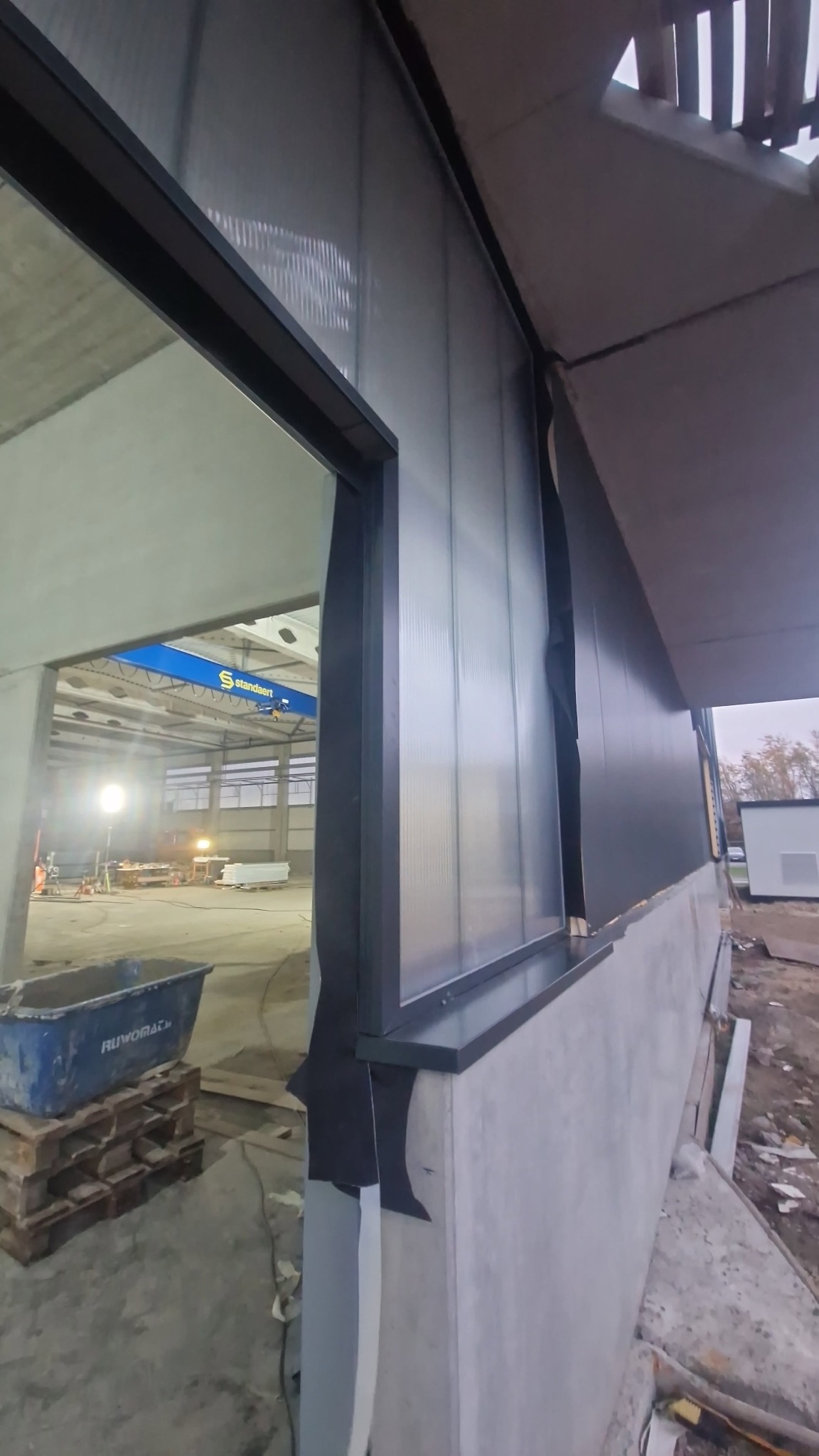 Montaż paneli warstwowych na hali, widok z zewnątrz. Widoczny fragment konstrukcji stalowej i wnętrze hali z suwnicą. Detal łączenia paneli.