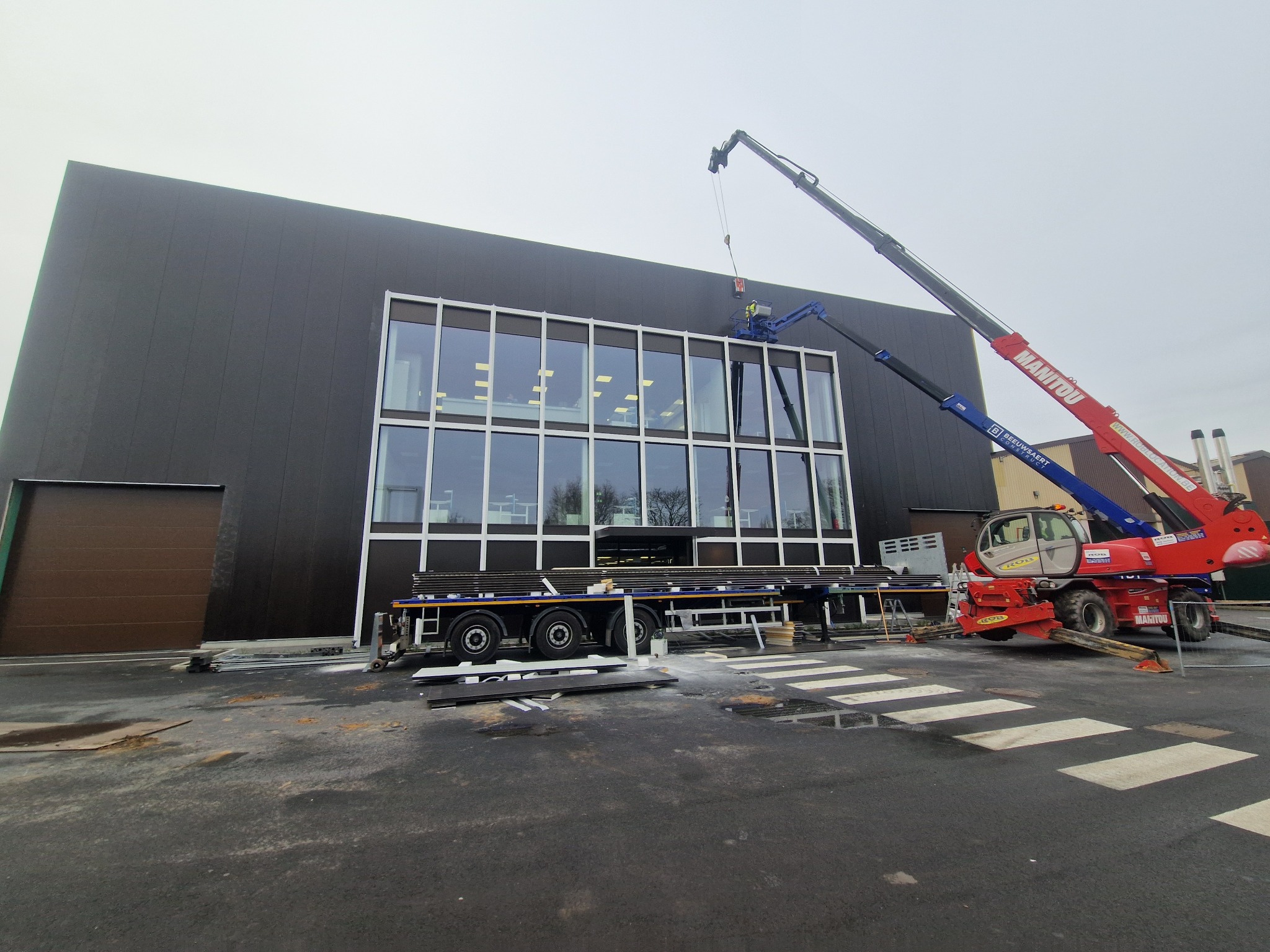 Montaż płyt warstwowych na hali z użyciem dźwigu Manitou. Widoczne płyty na przyczepie i pracownik w koszu dźwigu. Budynek z czarną elewacją i dużymi oknami.