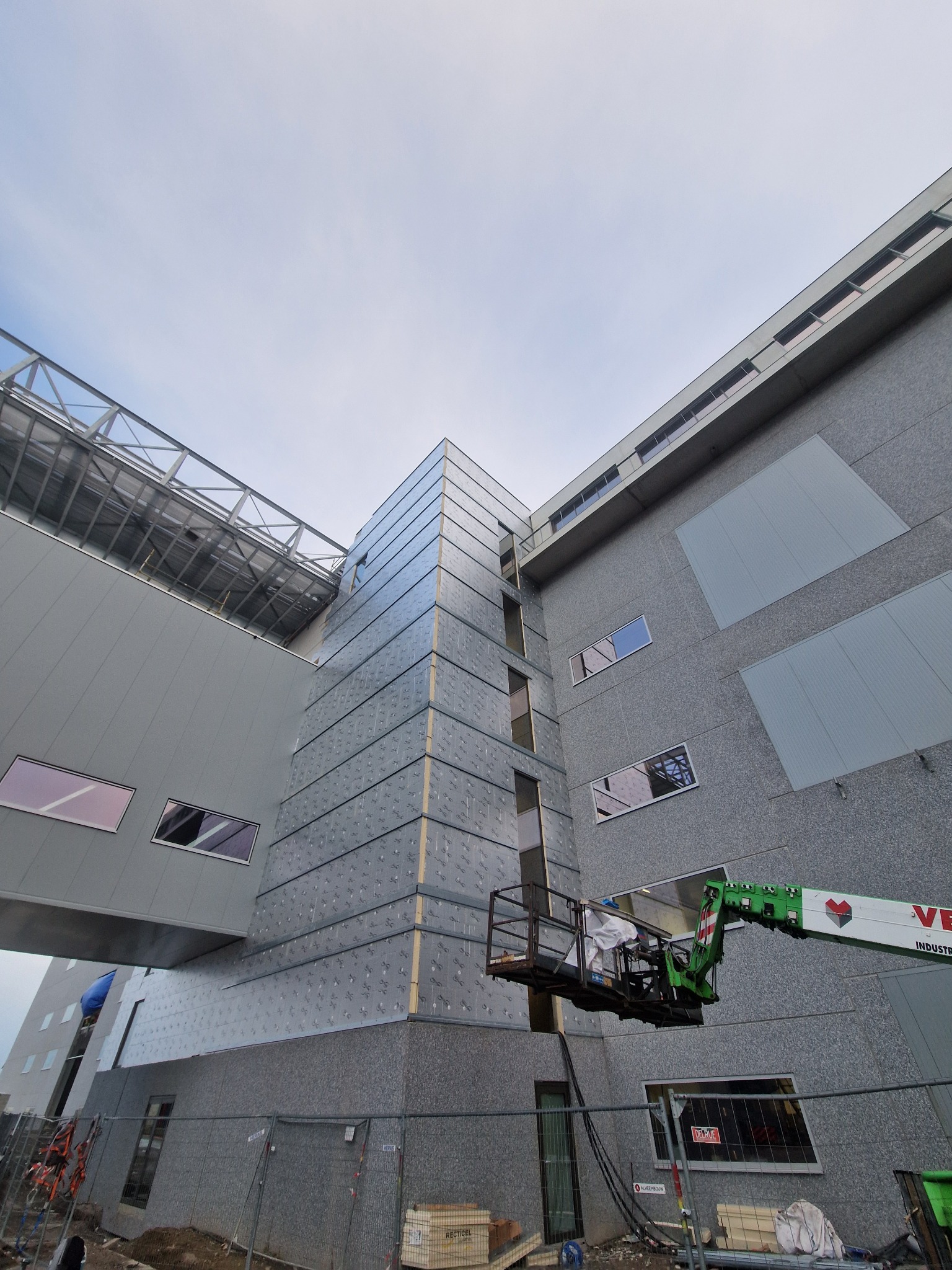 Montaż elewacji hali przemysłowej z podnośnika koszowego. Widoczne płyty warstwowe i elementy konstrukcyjne. Ujęcie z dołu, pochmurne niebo w tle.