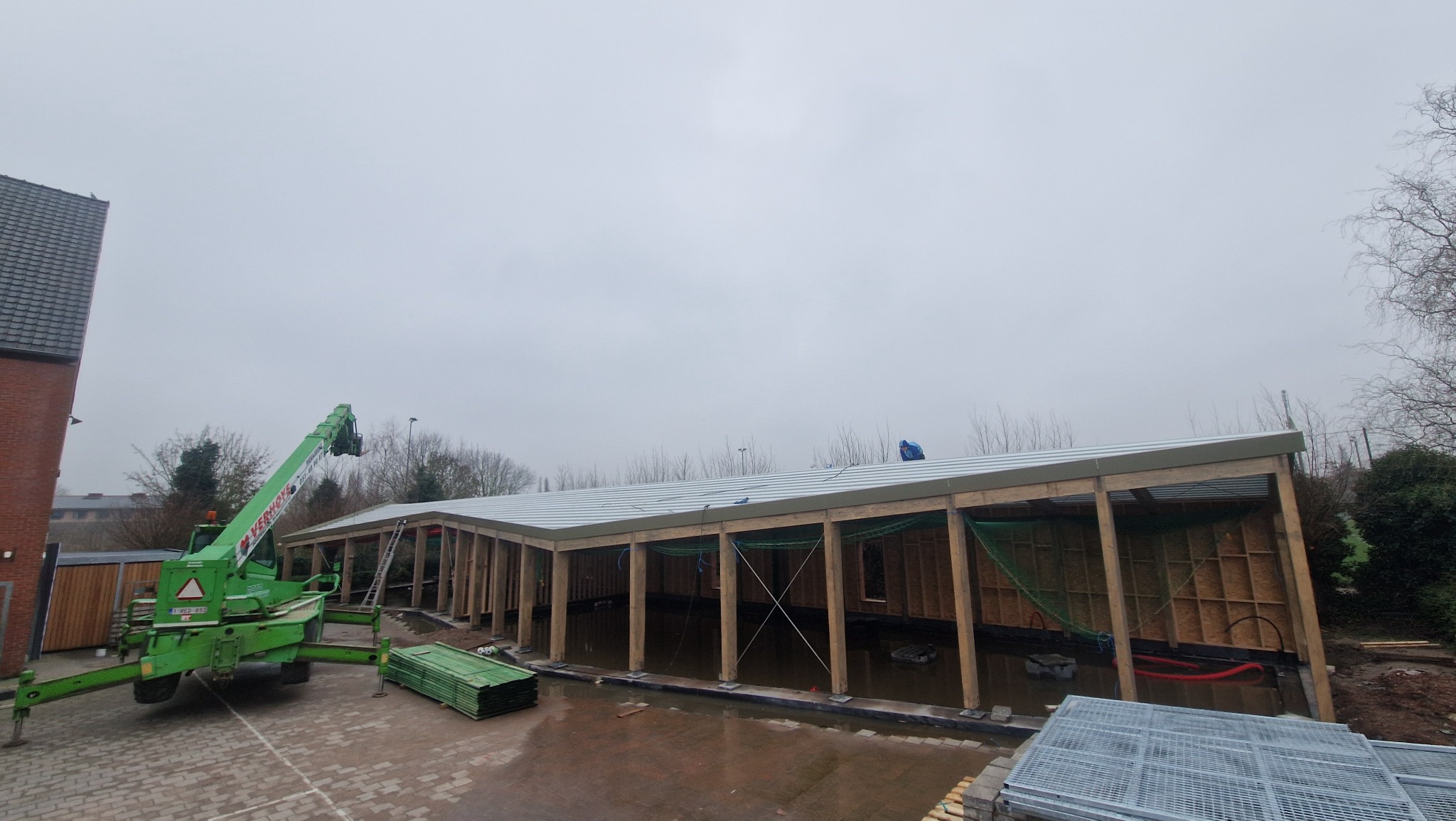 Budowa hali: drewniana konstrukcja z częściowo zamontowanym dachem z blachy trapezowej. Na pierwszym planie zielony dźwig i materiały budowlane.