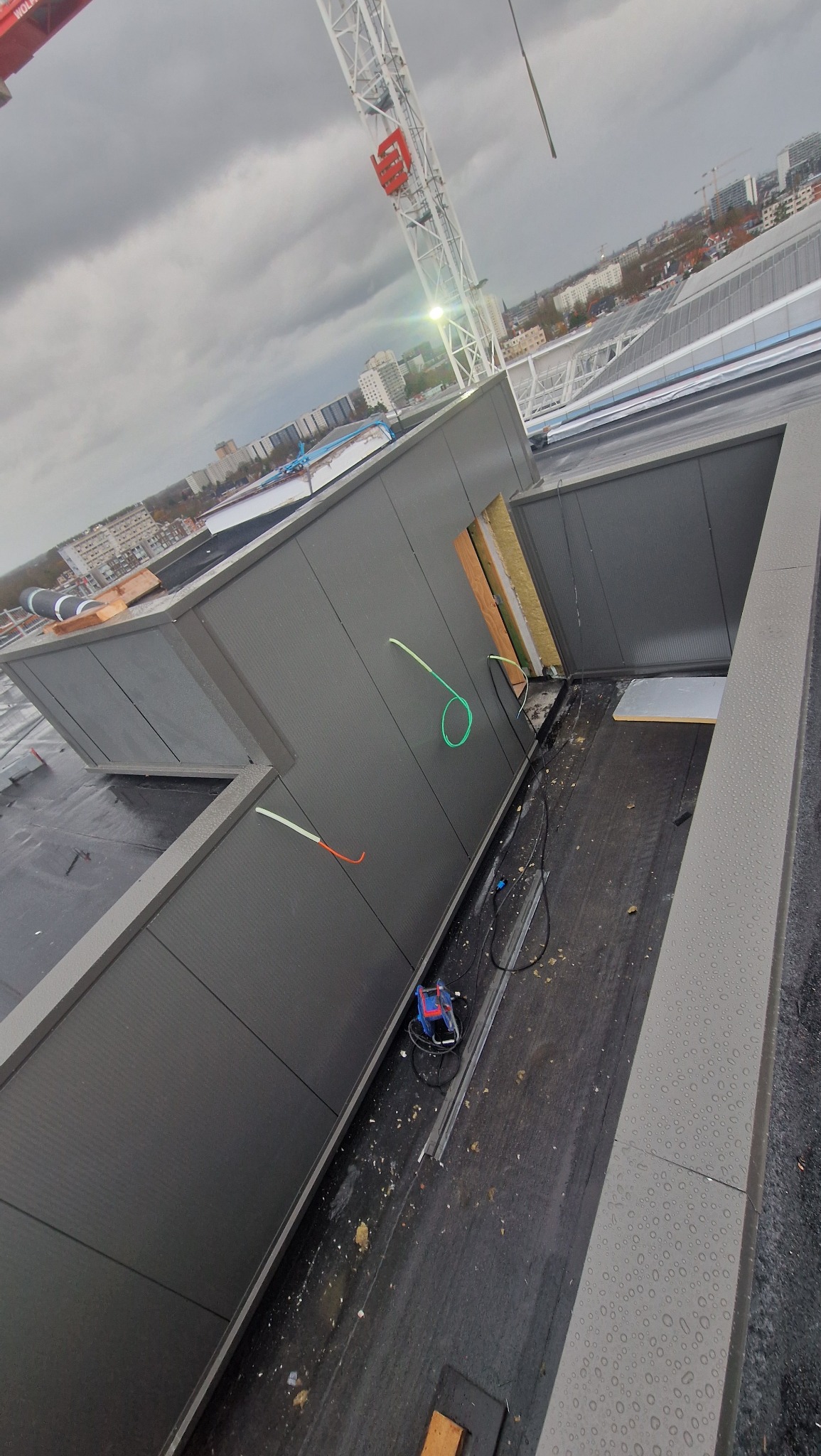 Montaż szarej płyty warstwowej na dachu budynku w trakcie budowy. Widoczny dźwig i elementy konstrukcyjne. Ujęcie z góry, pochmurne niebo w tle.