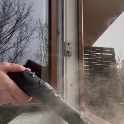 Ogarnij To - Czyszczenie parowe ramy okiennej z użyciem parownicy. Widoczna para wydobywająca się z dyszy urządzenia. Okno z widokiem na drzewa i balkon w tle.