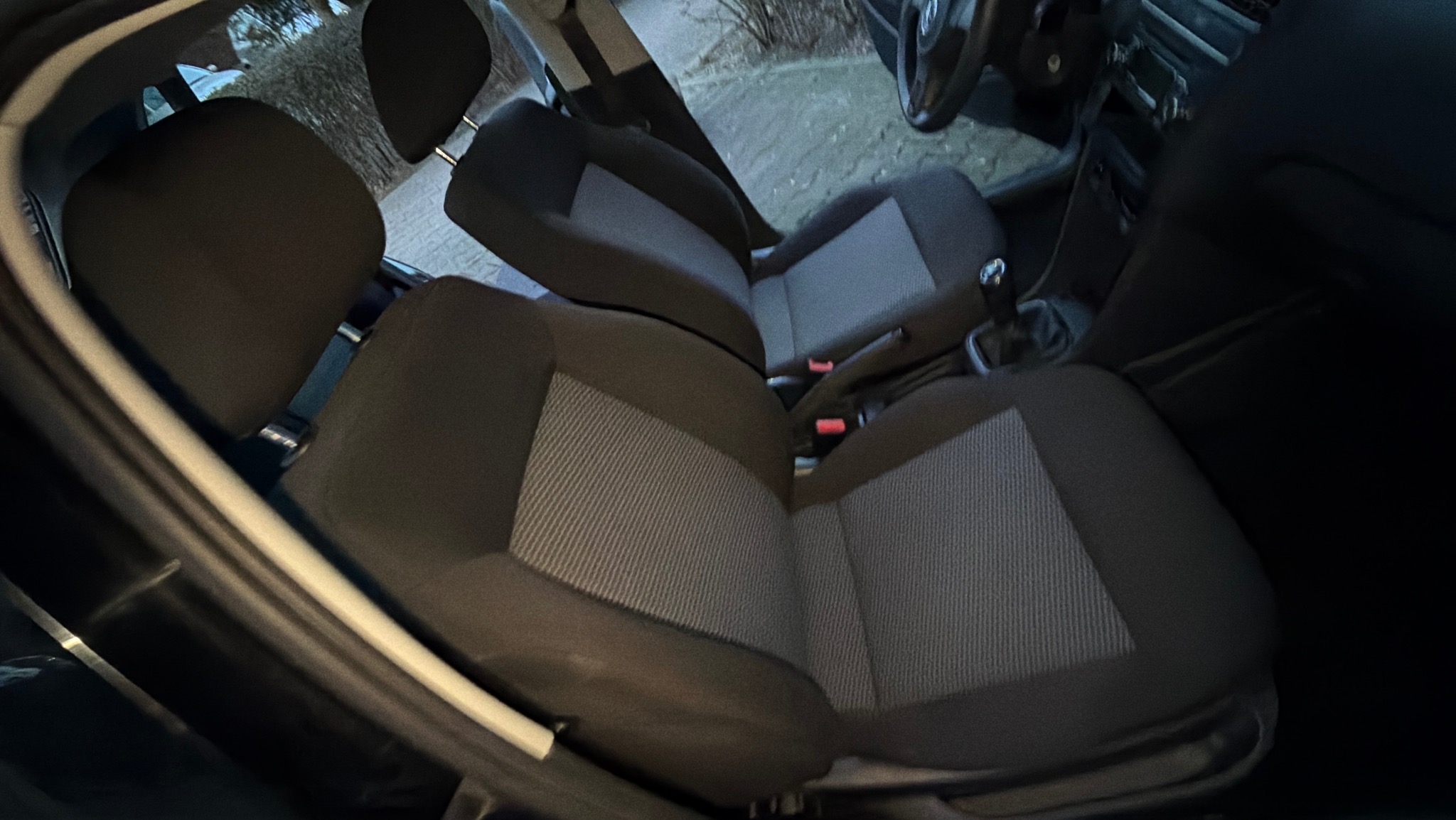 Wnętrze samochodu z widocznymi dwoma rzędami siedzeń z szarą tapicerką. Widoczna dźwignia zmiany biegów i fragment deski rozdzielczej. Ujęcie z perspektywy pasażera.