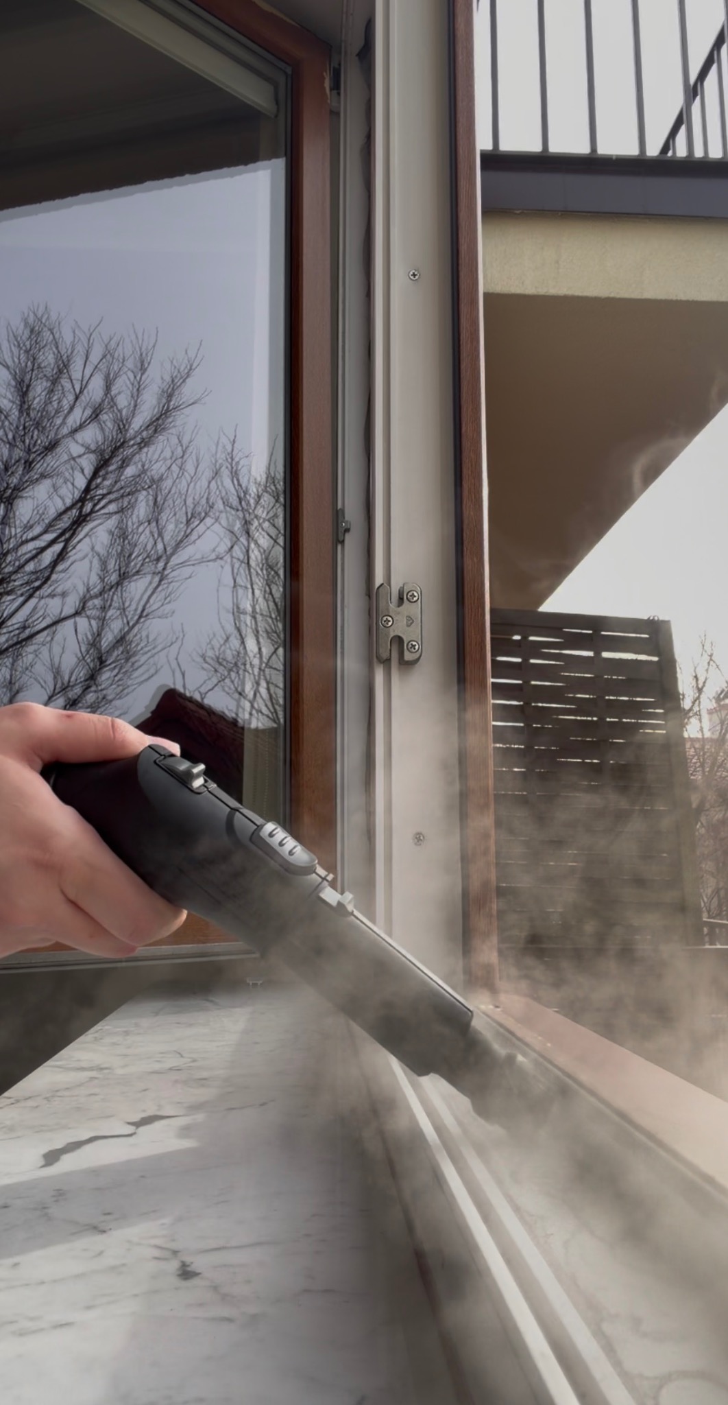 Czyszczenie parowe ramy okiennej z użyciem parownicy. Widoczna para wydobywająca się z dyszy urządzenia. Okno z widokiem na drzewa i balkon w tle.