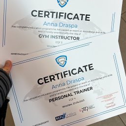 Anna Draspa - Dwa certyfikaty ukończenia kursów: instruktora siłowni i trenera personalnego, podpisane przez Annę Draspę, z logo organizacji certyfikujących. Ujęcie z bliska, lekki kąt.