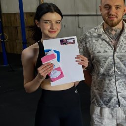 Anna Draspa - Uśmiechnięta kobieta w sportowym stroju trzymająca prezent (skarpety) i certyfikat z wizerunkiem trenera, obok stoi mężczyzna z tatuażem na szyi.