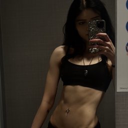 Anna Draspa - Kobieta w czarnym sportowym topie robi selfie, prezentując wyrzeźbiony brzuch i delikatny piercing pępka na tle szarej ściany. Widoczny cień i oświetlenie podkreślają definicję mięśni.