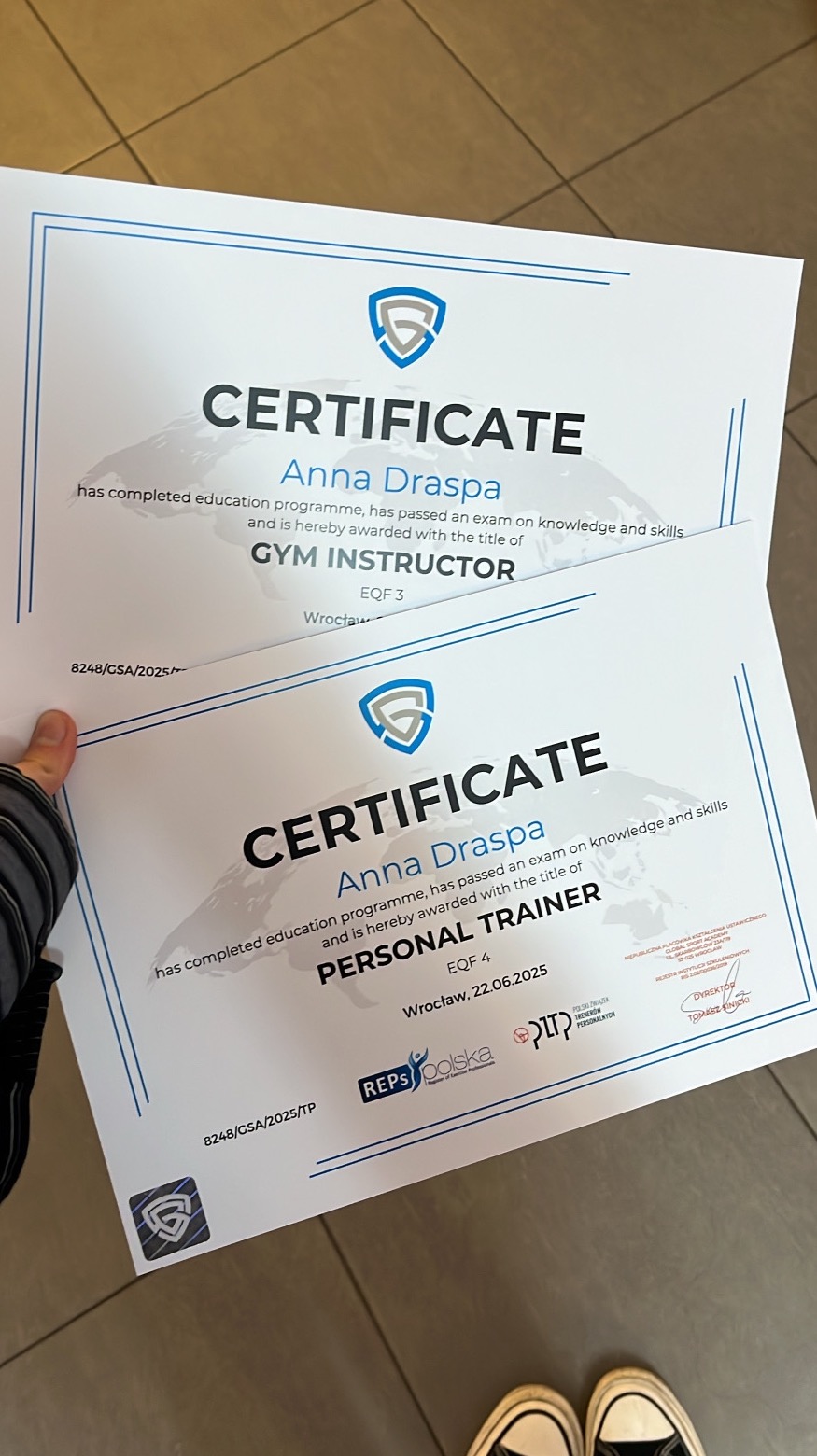 Dwa certyfikaty ukończenia kursów: instruktora siłowni i trenera personalnego, podpisane przez Annę Draspę, z logo organizacji certyfikujących. Ujęcie z bliska, lekki kąt.