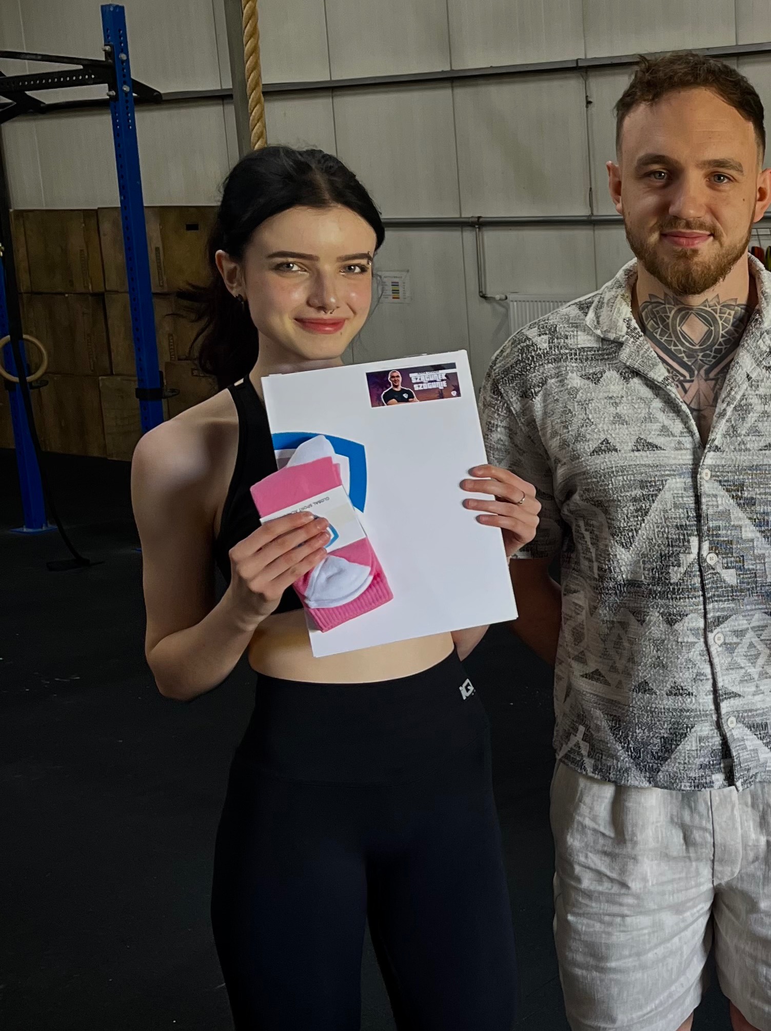 Uśmiechnięta kobieta w sportowym stroju trzymająca prezent (skarpety) i certyfikat z wizerunkiem trenera, obok stoi mężczyzna z tatuażem na szyi.