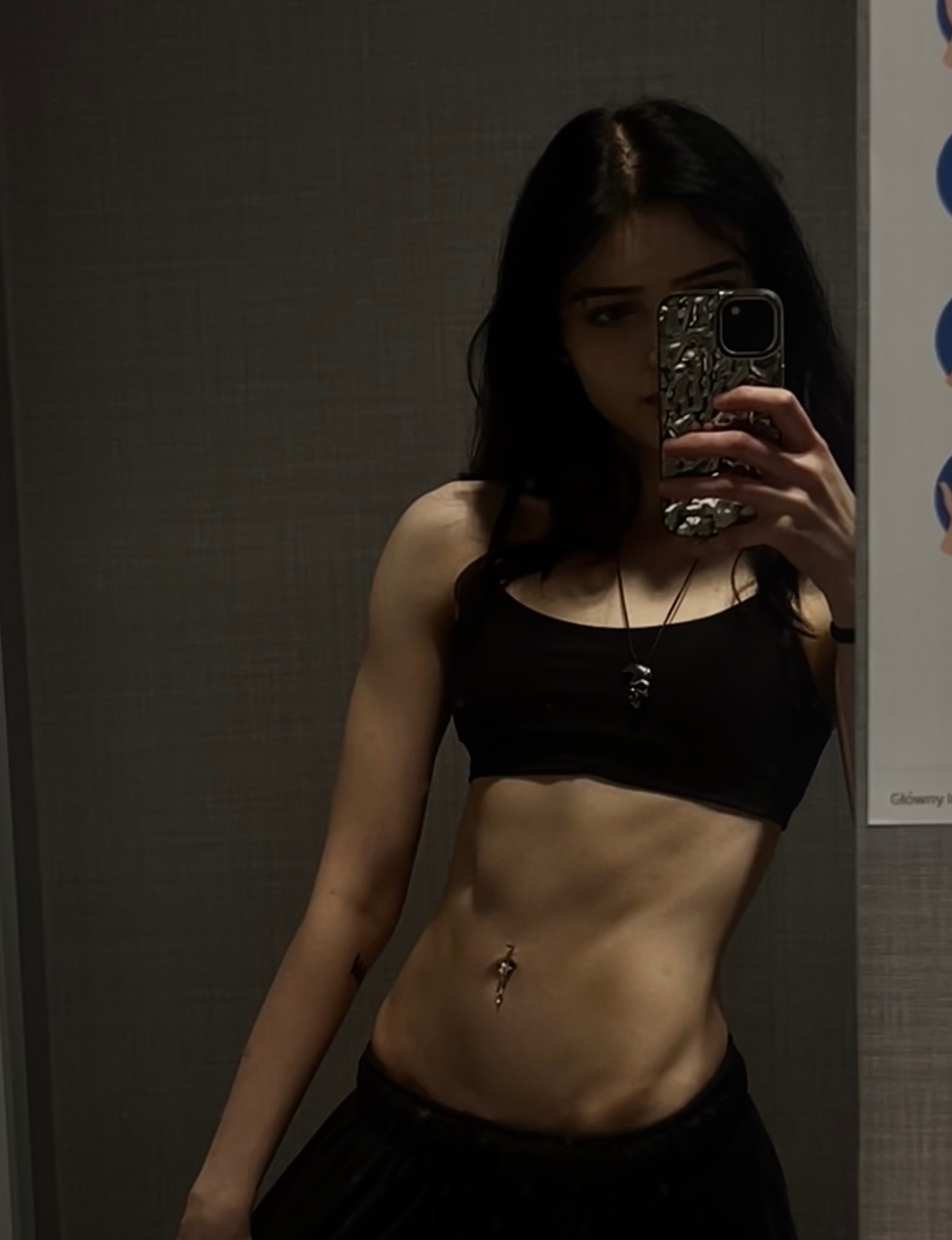 Kobieta w czarnym sportowym topie robi selfie, prezentując wyrzeźbiony brzuch i delikatny piercing pępka na tle szarej ściany. Widoczny cień i oświetlenie podkreślają definicję mięśni.