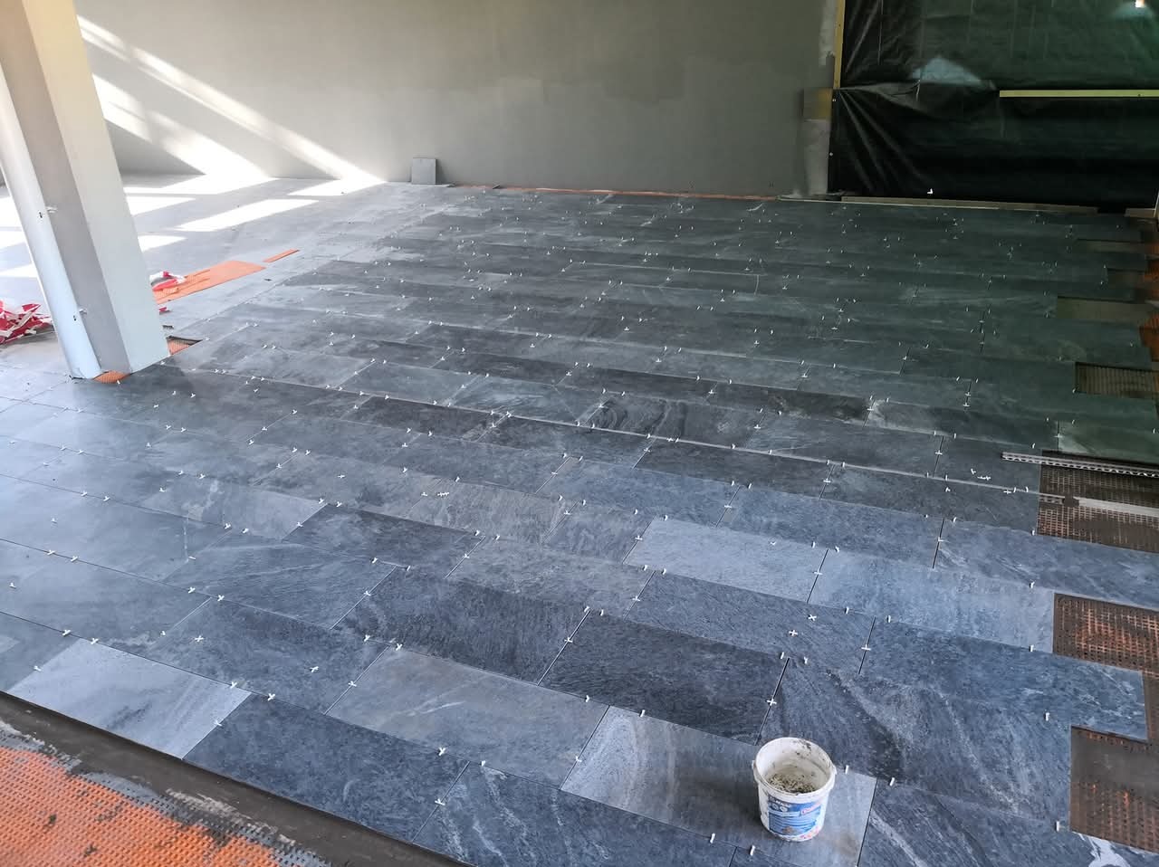 Układanie podłogi z płytek imitujących kamień, z użyciem krzyżyków dystansowych. Widoczny fragment ściany i kolumna. Wiaderko z klejem w rogu kadru.