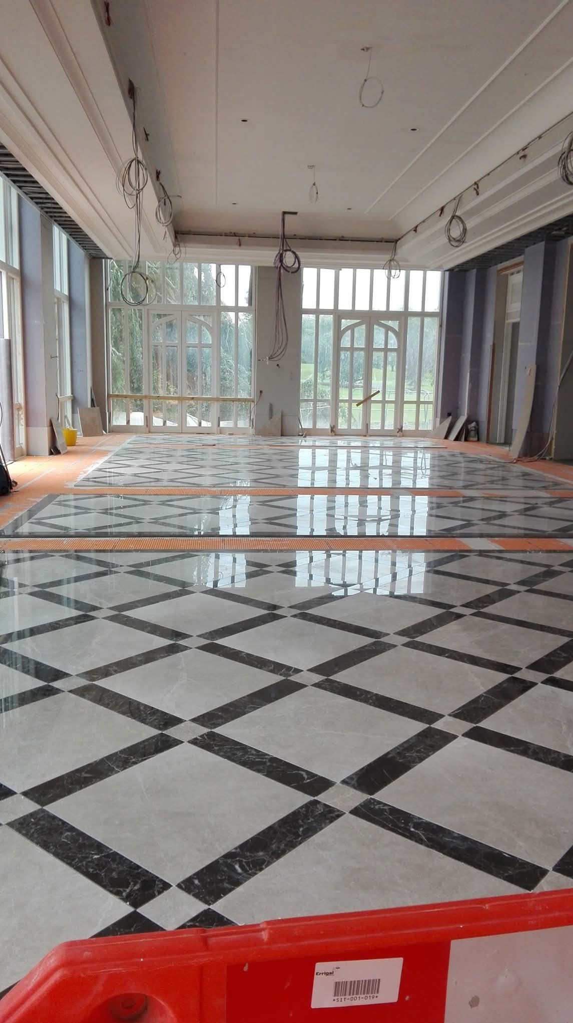 Ułożona podłoga z płytek w szachownicę w przestronnym, jasnym pomieszczeniu z dużymi oknami. Widoczne przygotowania do dalszych prac wykończeniowych.