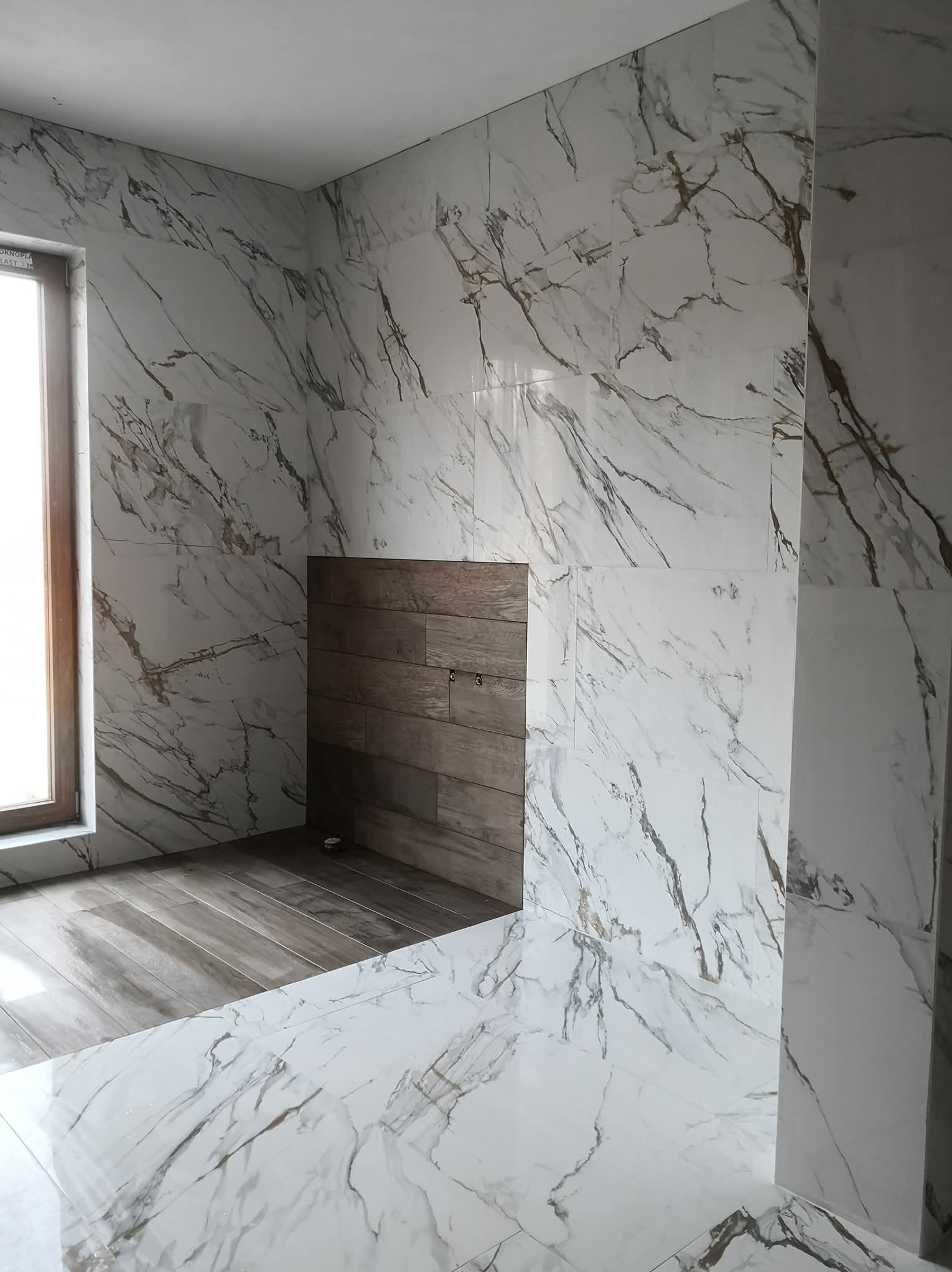 Wykończona łazienka z białymi płytkami imitującymi marmur z brązowymi żyłami na ścianach i podłodze, oraz drewnopodobnymi płytkami na fragmencie ściany.