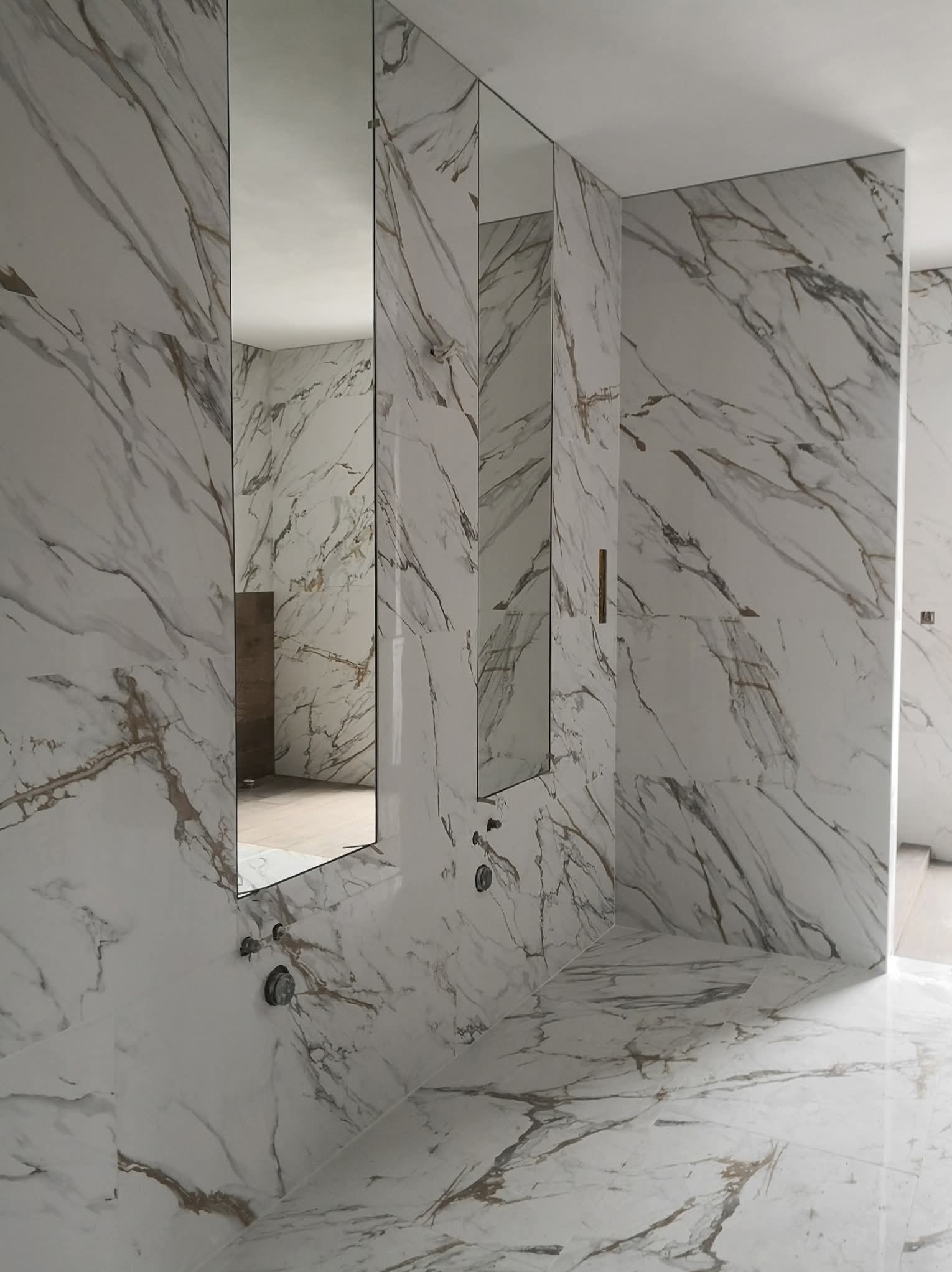 Wykończona łazienka z białymi płytkami imitującymi marmur, z lustrami w wąskich ramach. Widoczne elementy armatury i fragment podłogi. Minimalistyczny design.