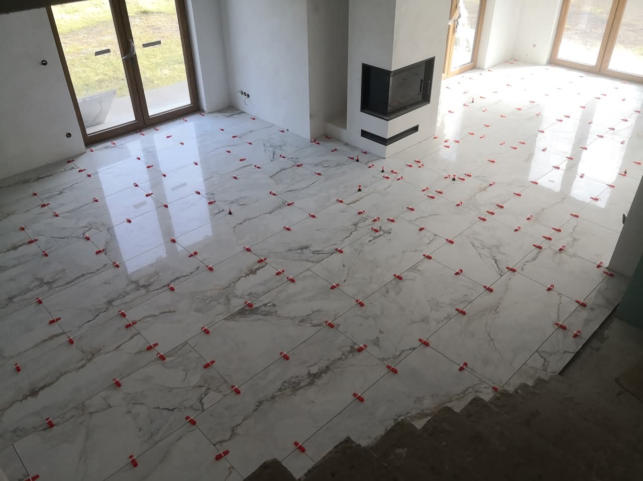 Układanie marmurowych płytek podłogowych w salonie z kominkiem, z użyciem czerwonych klipsów poziomujących. Widoczne okna i schody.