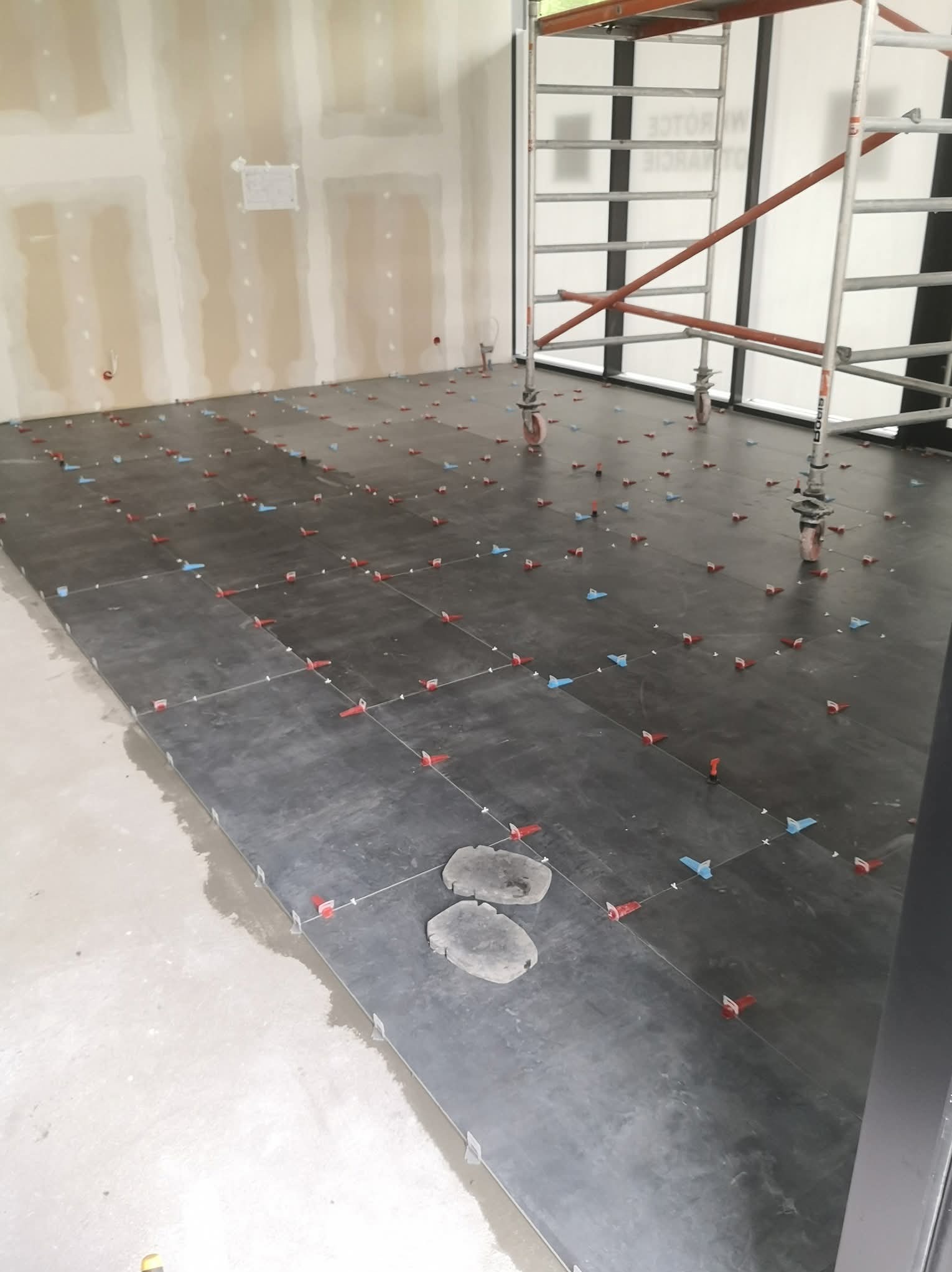 Układanie ciemnych płytek podłogowych z użyciem systemu poziomowania, widoczne krzyżyki i kliny. W tle ściany przygotowane do malowania i rusztowanie.