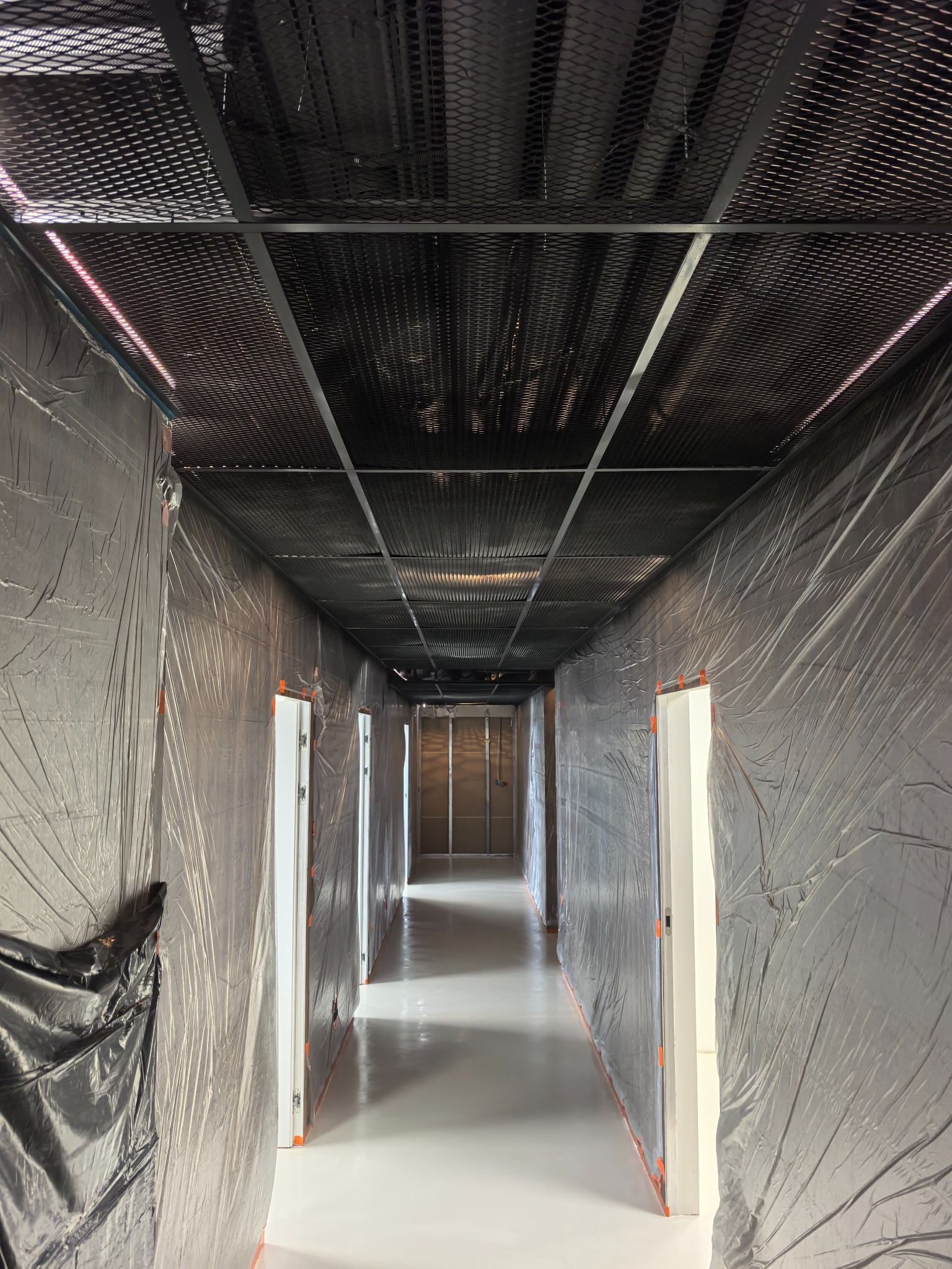 Korytarz w trakcie remontu: podłoga zabezpieczona folią, ściany osłonięte, sufit z metalowej siatki. Widoczne białe drzwi i przygotowania do dalszych prac wykończeniowych.