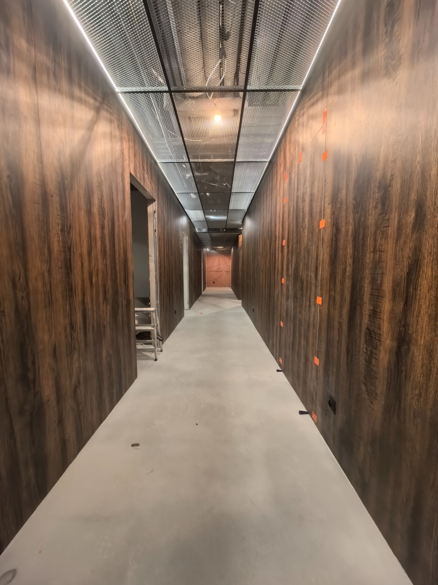 Korytarz w trakcie wykończenia: drewnopodobne panele ścienne, sufit z metalowej siatki z oświetleniem LED, szpachlowana podłoga, drabina w tle.