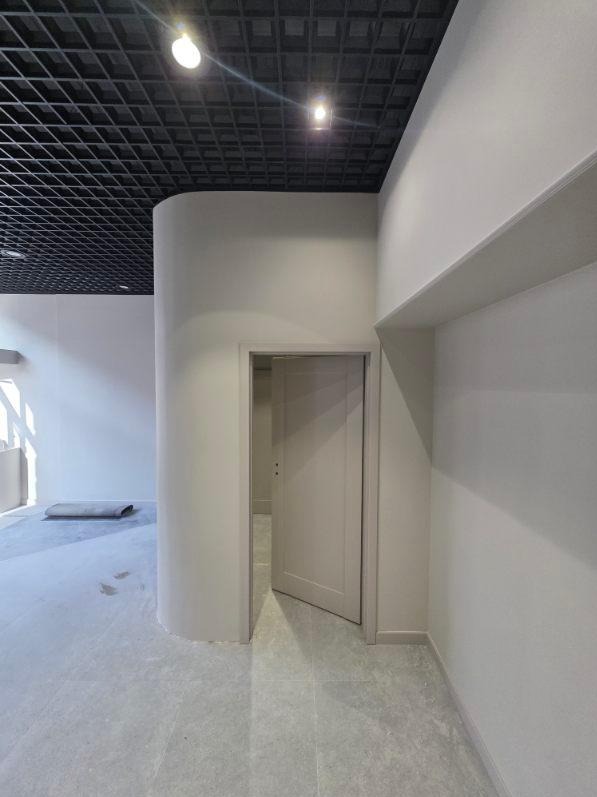 Jasne wnętrze z otwartymi drzwiami i zaokrąglonym narożnikiem ściany. Czarna, kratowa struktura sufitu. Minimalistyczny design, neutralna kolorystyka.