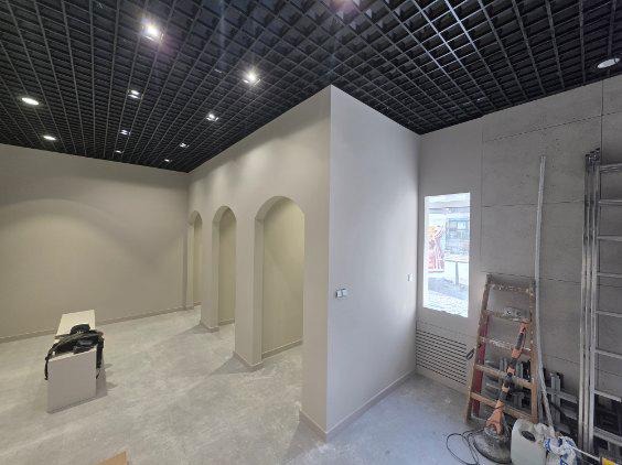 Wnętrze w trakcie prac wykończeniowych: szara podłoga, łukowe przejścia, czarny sufit kasetonowy z oświetleniem. Widoczne drabiny i narzędzia.