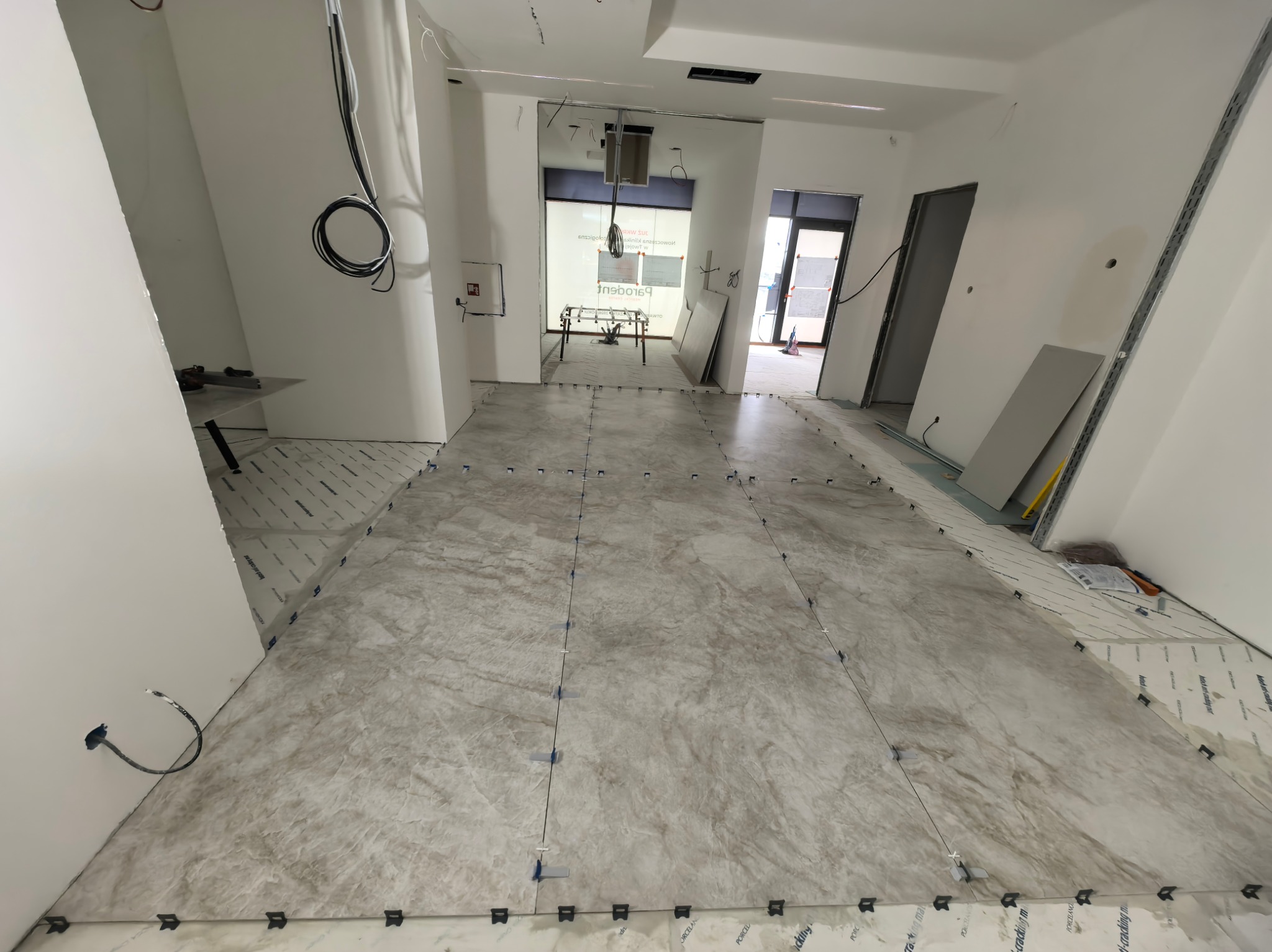 Układanie płytek podłogowych w jasnym wnętrzu z widocznymi krzyżykami dystansowymi i folią ochronną. Prace glazurnicze w trakcie realizacji.