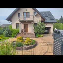 WIBRUK - Dom jednorodzinny z podjazdem wyłożonym kostką w odcieniach beżu, otoczony zielenią i ogrodzeniem panelowym. Na dachu panele słoneczne.