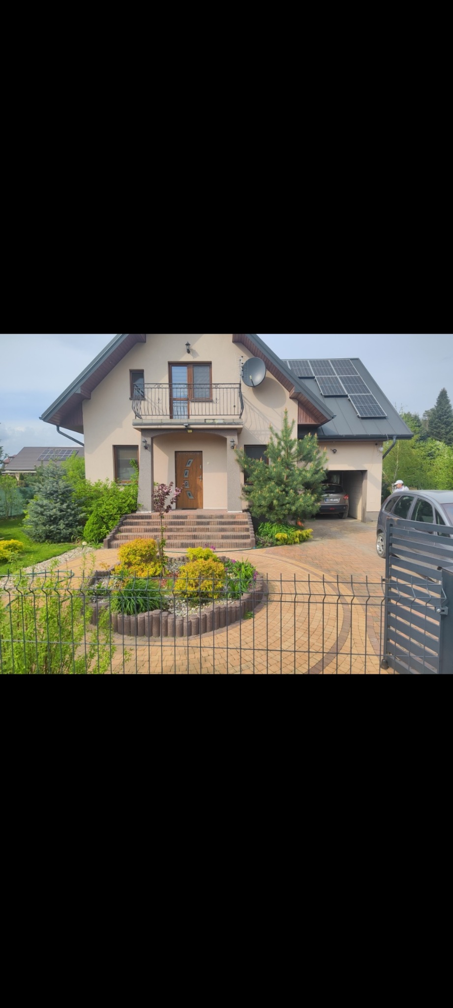 Dom jednorodzinny z podjazdem wyłożonym kostką w odcieniach beżu, otoczony zielenią i ogrodzeniem panelowym. Na dachu panele słoneczne.