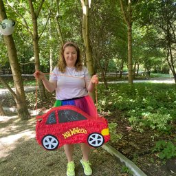 FANIMACJE Patrycja Jankowska - Animator w lesie trzyma czerwoną piniatę w kształcie auta Hot Wheels z imionami dzieci. Kolorowa spódnica, drzewa w tle, balon z pandą.