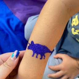 FANIMACJE Patrycja Jankowska - Błyszczący, fioletowy tatuaż z dinozaurem na ramieniu dziecka, wykonany podczas imprezy. Widoczna dłoń z pomalowanymi paznokciami i kolorowe tło.
