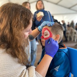 FANIMACJE Patrycja Jankowska - Malowanie twarzy chłopca na motyw Spidermana przez kobietę w niebieskich rękawiczkach, w tle inne osoby w namiocie.