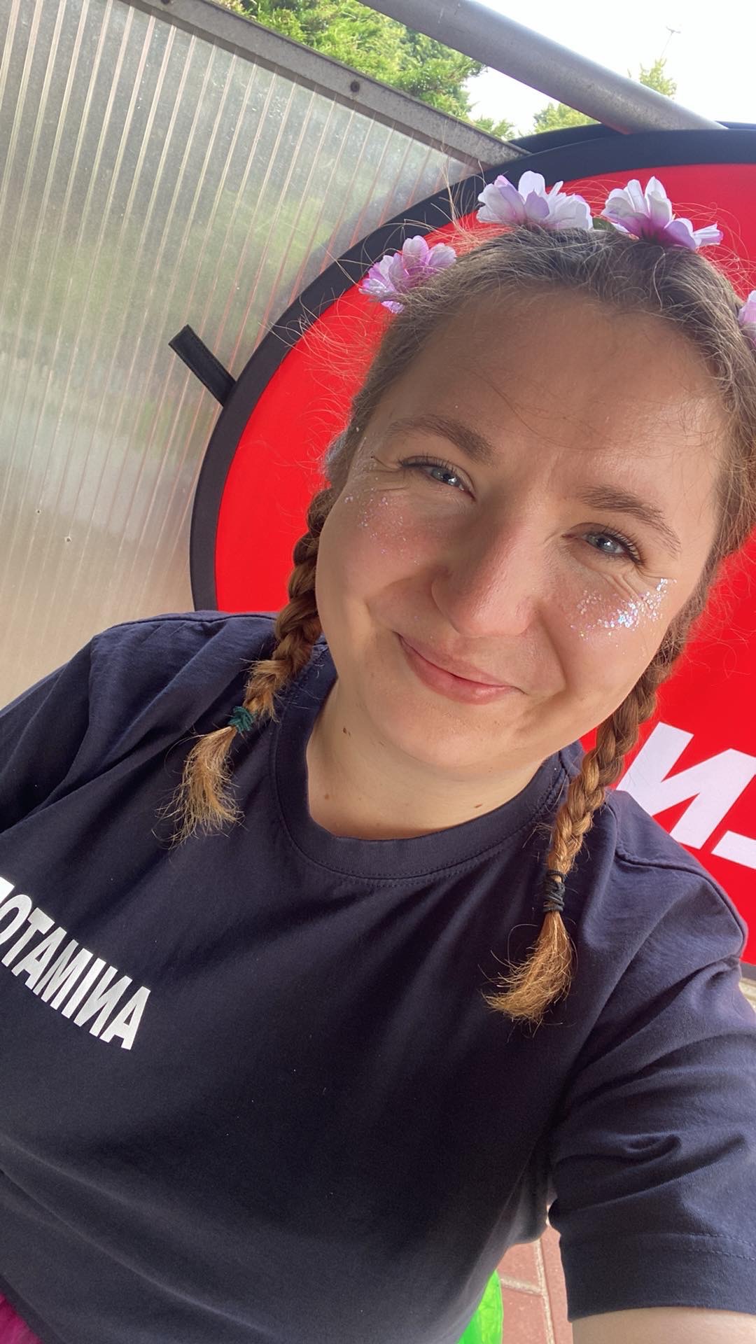 Uśmiechnięta kobieta z brokatem na twarzy i wiankiem z kwiatów na głowie, w tle czerwony element dekoracyjny. Włosy zaplecione w dwa warkocze.