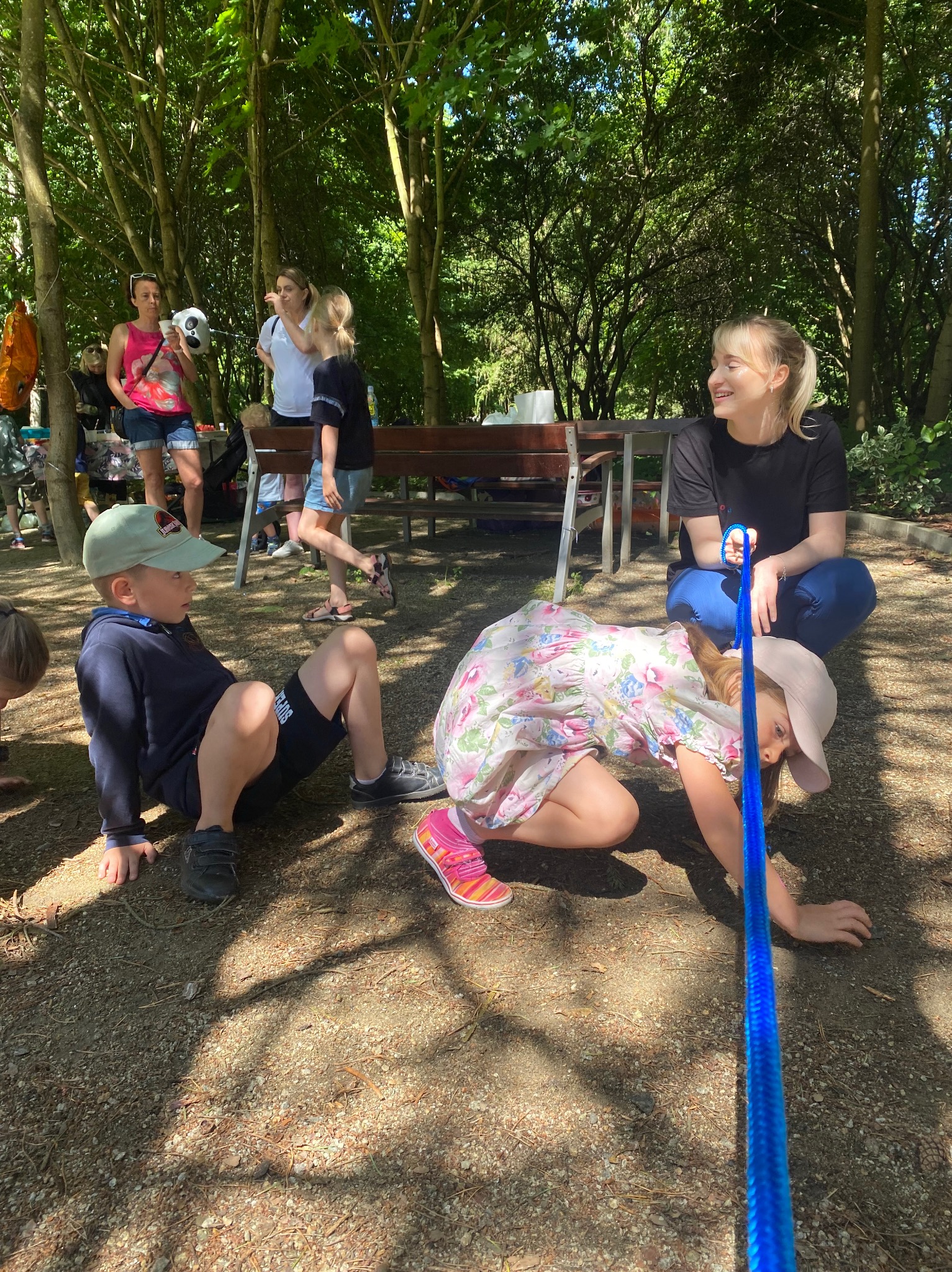 Animator prowadzi zabawę z dziećmi na świeżym powietrzu. Dziewczynka w kapeluszu i sukience na czworakach, chłopiec siedzi obok. W tle inne dzieci i opiekunowie.