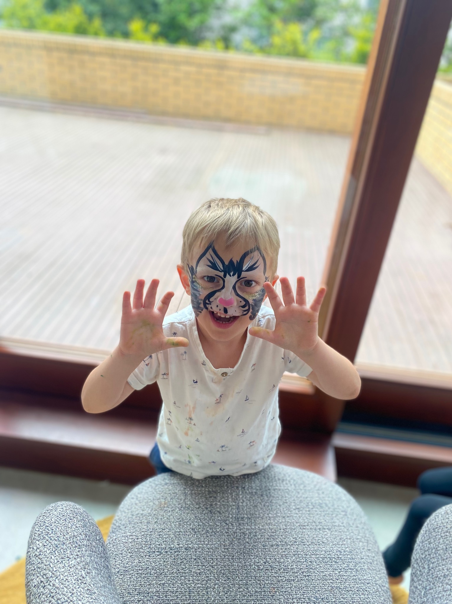 Uśmiechnięty chłopiec z pomalowaną twarzą w motywy zwierzęce, wyciąga ręce do przodu, siedząc na fotelu w jasnym wnętrzu z dużym oknem.