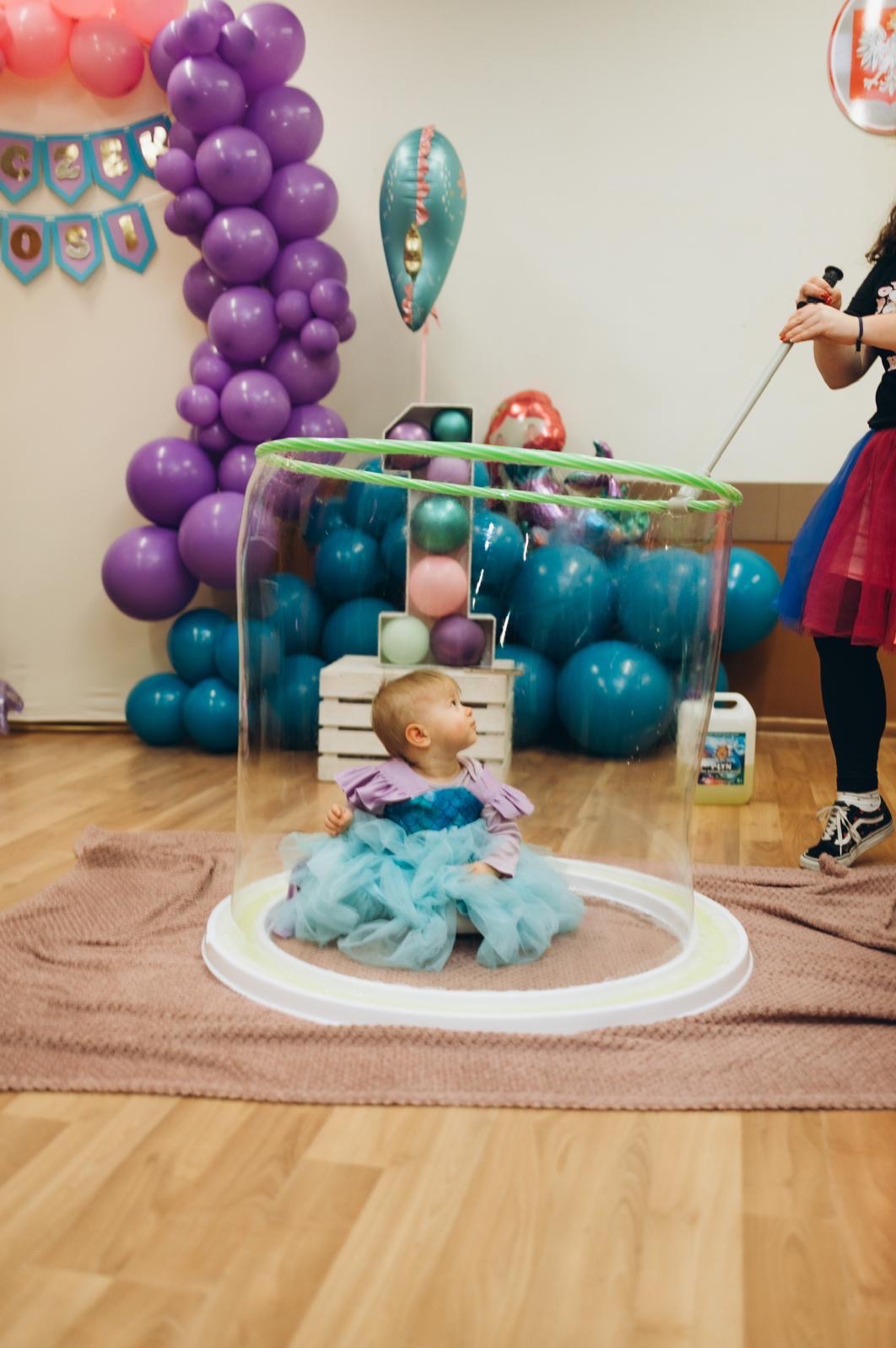 Dziewczynka w błękitnej sukience siedzi wewnątrz dużej bańki mydlanej, otoczona dekoracjami balonowymi na przyjęciu urodzinowym. Widoczna osoba obsługująca bańki.