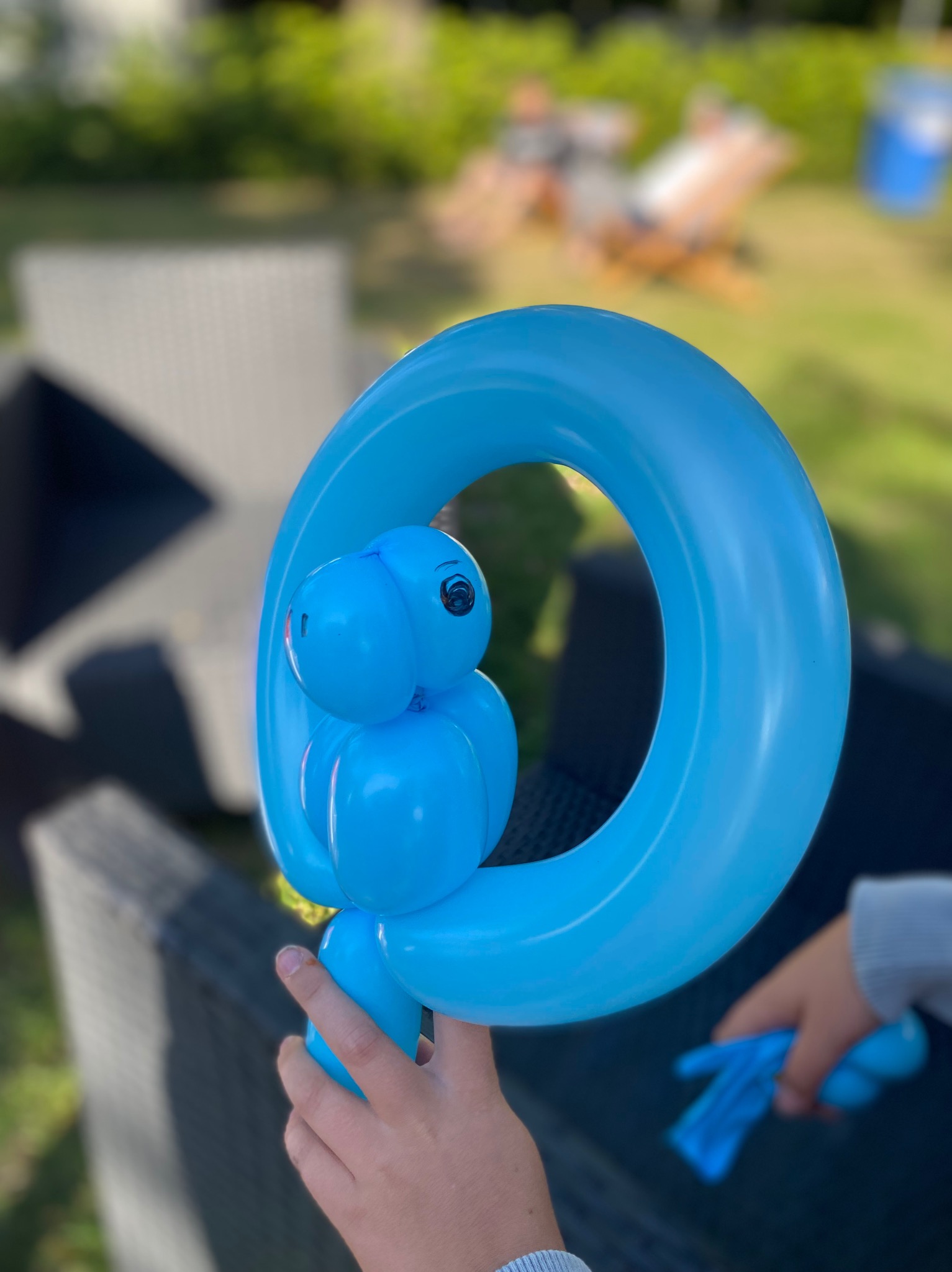 Niebieski balon w kształcie dinozaura trzymany przez dziecko na tle ogrodu. Balonowa figurka z okrągłym korpusem i małą głową.