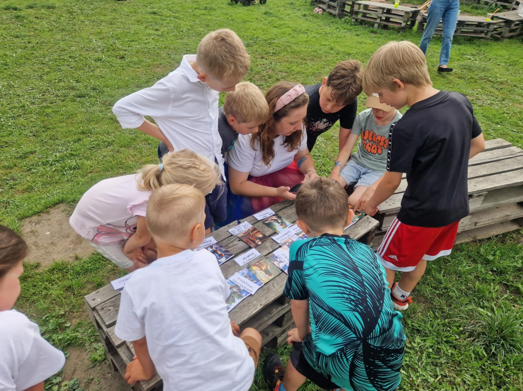 Grupa dzieci otacza kobietę przy drewnianym stole, oglądając karty z postaciami, na trawiastym terenie. Uczestnicy w różnorodnych strojach, skupieni na grze.