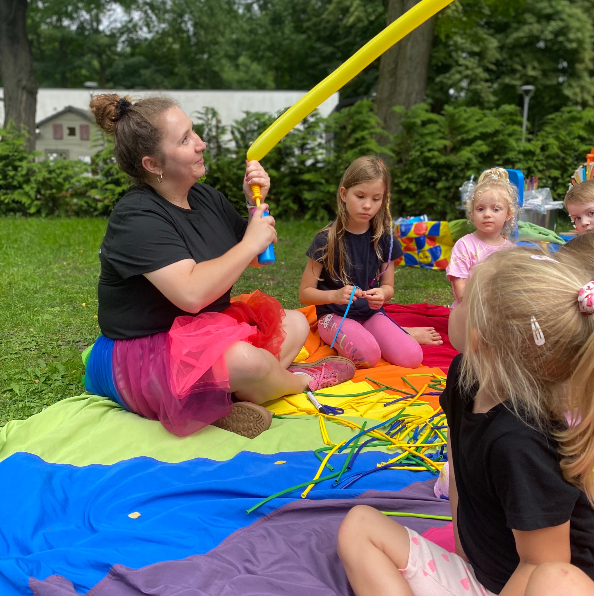 Animator w kolorowej spódnicy modeluje balon dla dzieci siedzących na tęczowym kocu podczas imprezy plenerowej. W tle zieleń drzew i budynek.