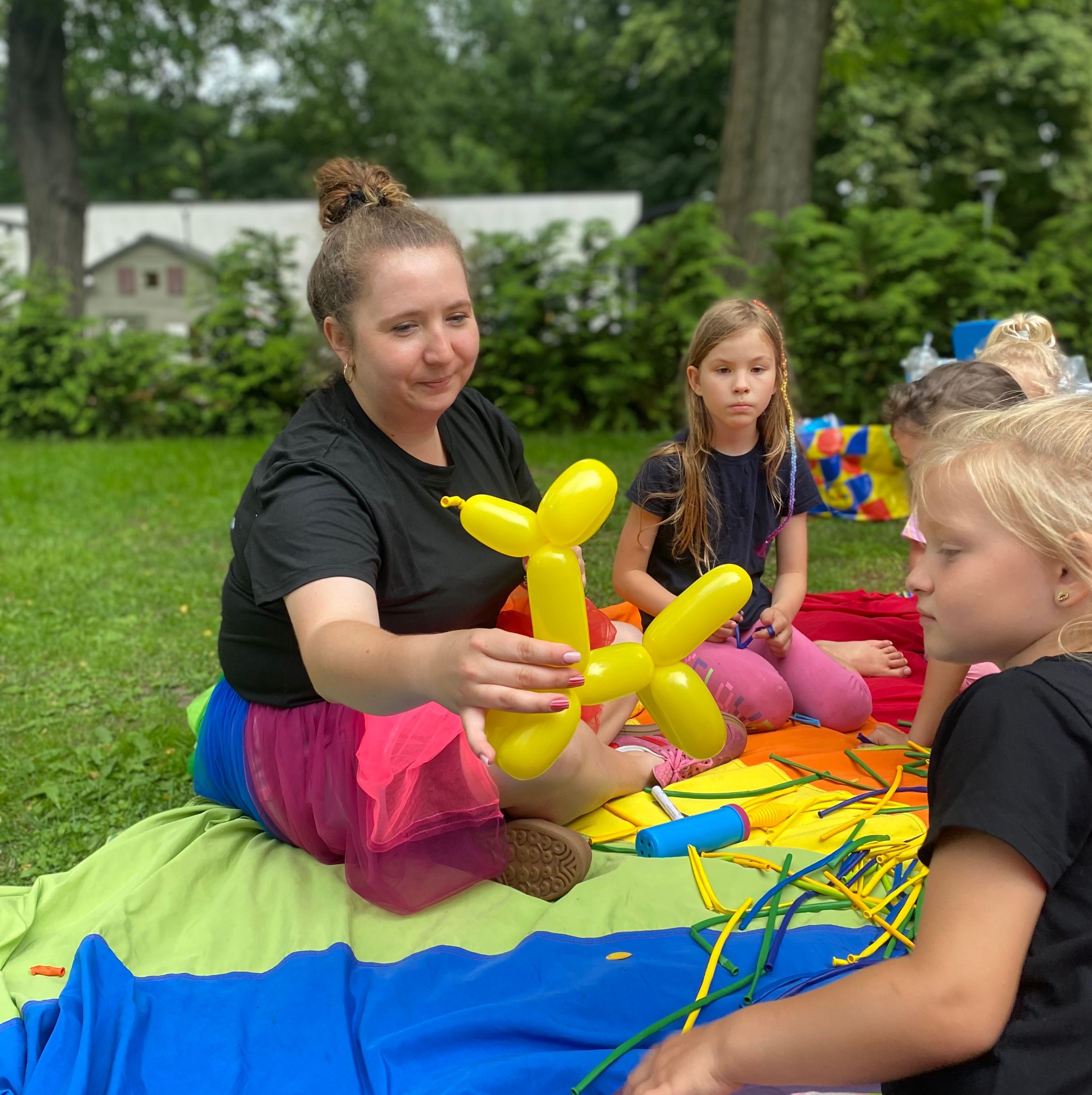 Animator w tęczowej spódnicy modeluje balon w kształcie psa dla dzieci siedzących na kocu na trawie. W tle drzewa i budynek.