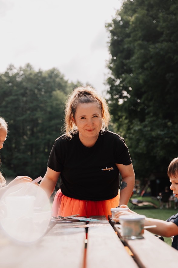 Uśmiechnięta animatorka w czarnym T-shircie i pomarańczowej tiulowej spódnicy prowadzi warsztaty plastyczne dla dzieci na świeżym powietrzu przy drewnianym stole.