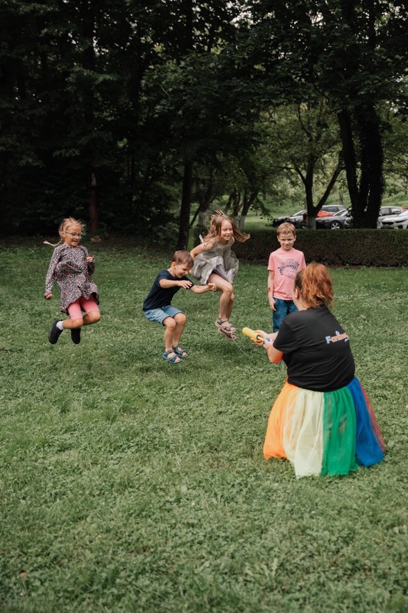 Animator w tęczowej spódnicy bawi grupę skaczących dzieci na zielonej trawie. W tle drzewa i zaparkowane samochody. Zabawa na świeżym powietrzu.