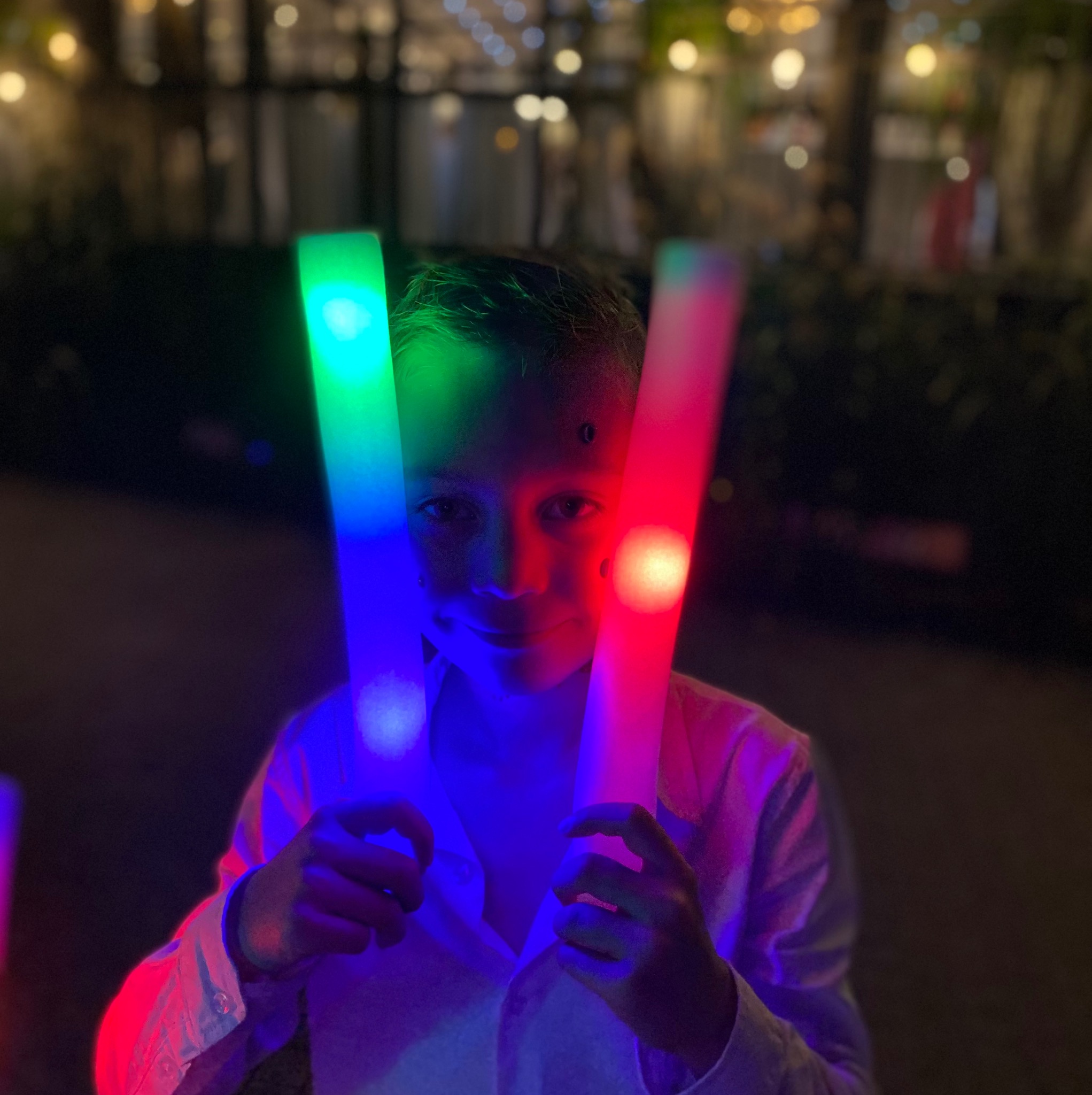 Uśmiechnięty chłopiec trzymający w dłoniach świecące, kolorowe pałeczki (czerwona i zielono-niebieska) na tle rozmytych światełek, prawdopodobnie podczas imprezy.