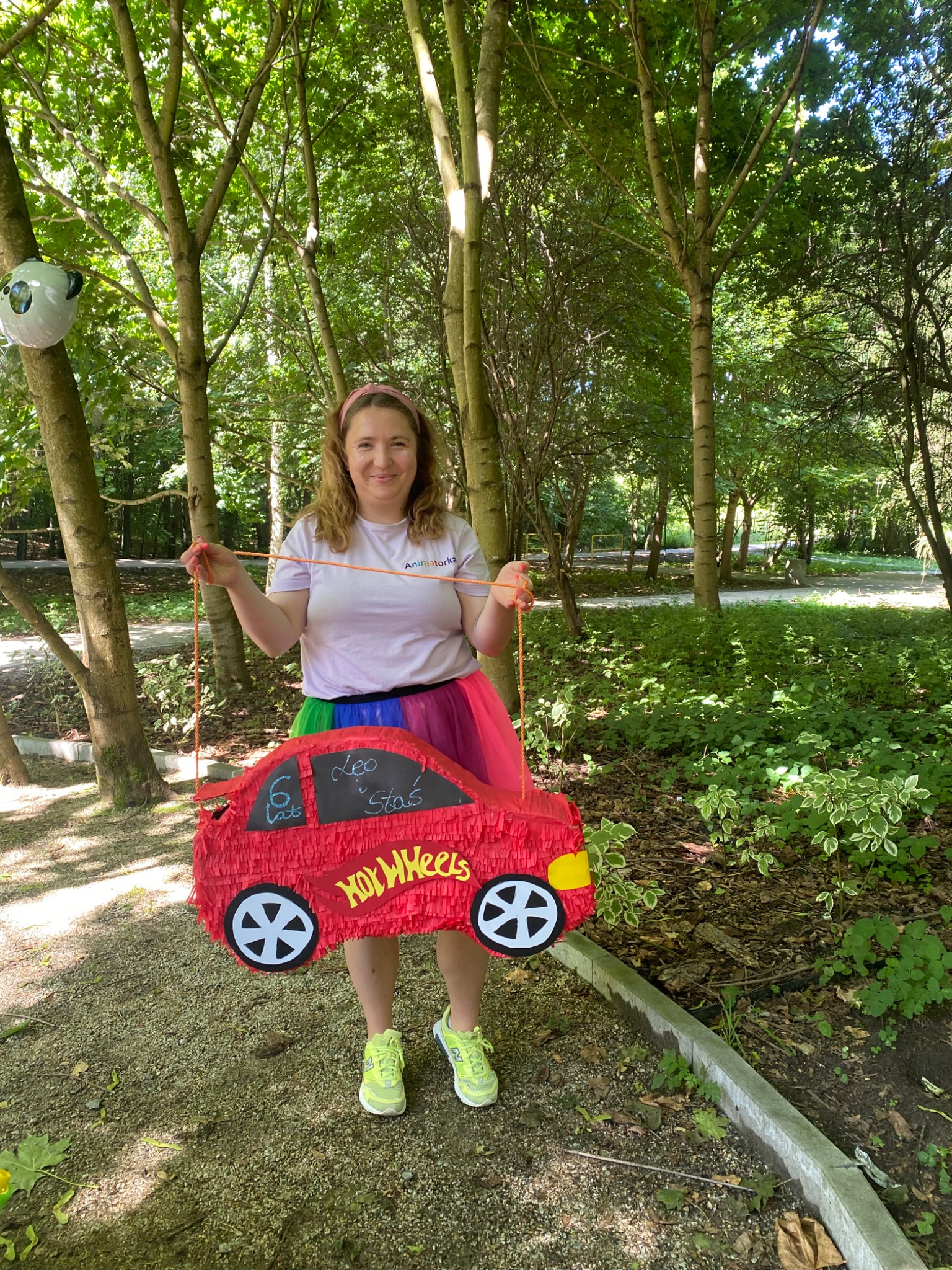 Animator w lesie trzyma czerwoną piniatę w kształcie auta Hot Wheels z imionami dzieci. Kolorowa spódnica, drzewa w tle, balon z pandą.