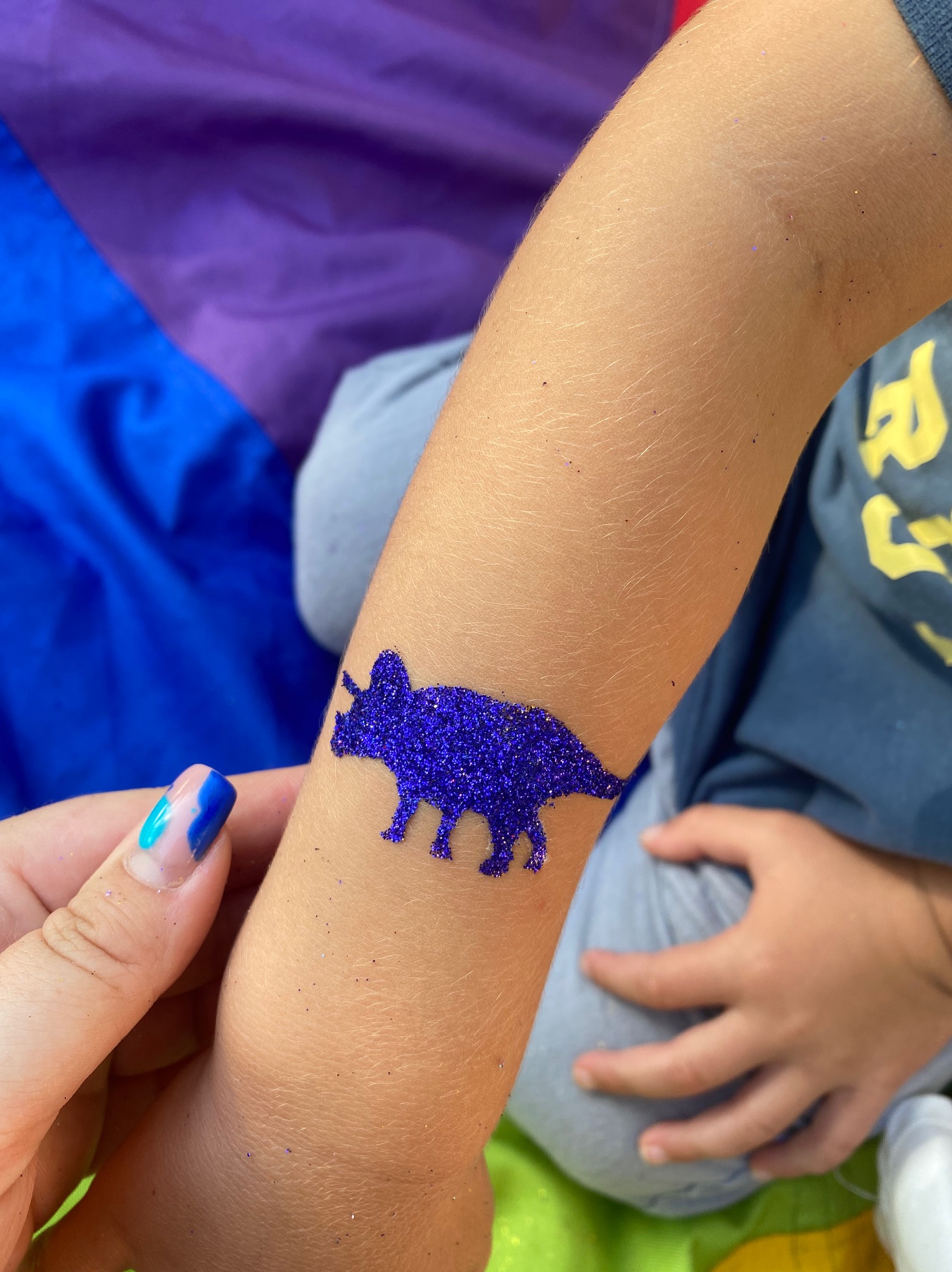 Błyszczący, fioletowy tatuaż z dinozaurem na ramieniu dziecka, wykonany podczas imprezy. Widoczna dłoń z pomalowanymi paznokciami i kolorowe tło.