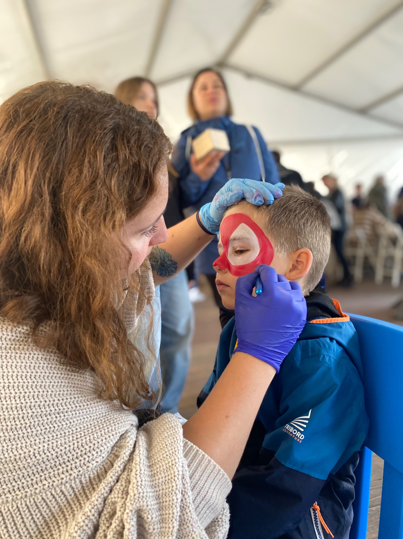 Malowanie twarzy chłopca na motyw Spidermana przez kobietę w niebieskich rękawiczkach, w tle inne osoby w namiocie.