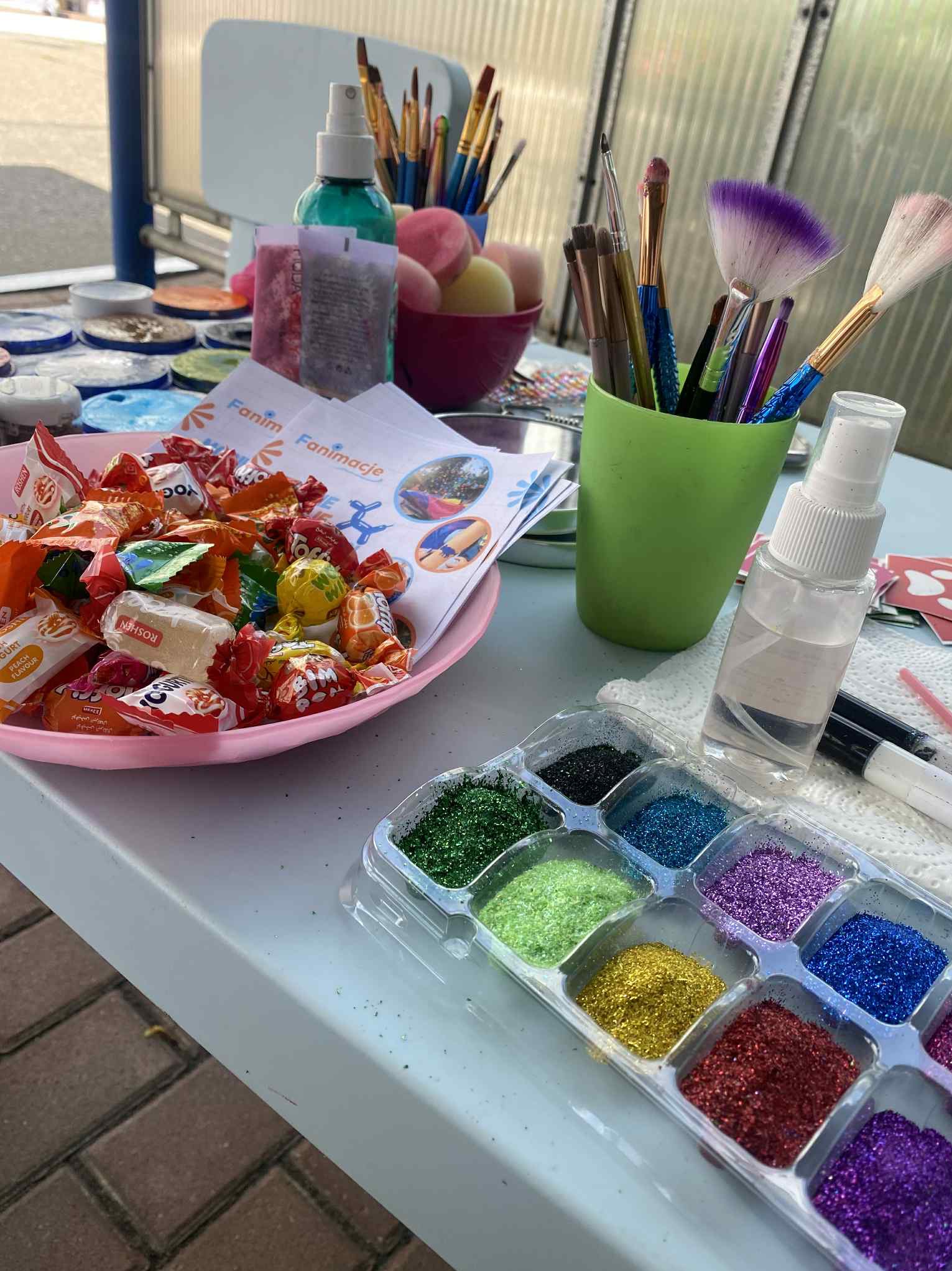 Stół z akcesoriami do malowania twarzy: pędzle, brokat, farby, słodycze i ulotki informacyjne. Przygotowania do animacji dla dzieci.