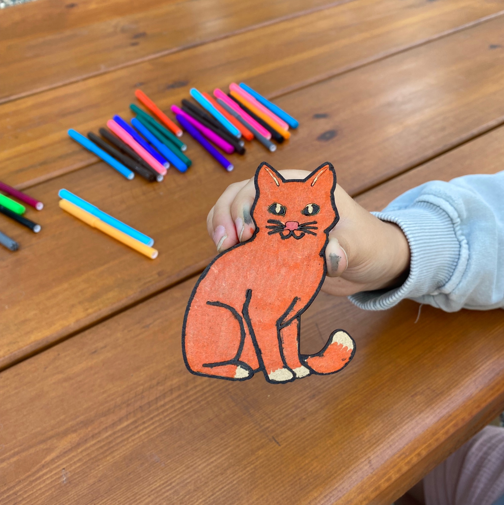 Dziecko trzyma pomalowaną na pomarańczowo figurkę kota na drewnianym stole, obok leżą kolorowe pisaki. Ręka dziecka jest lekko ubrudzona farbą.