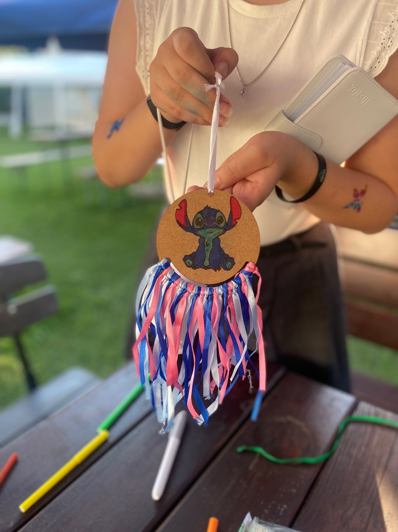 Osoba trzyma ręcznie robiony, okrągły brelok z postacią Stitcha i kolorowymi wstążkami, na tle stołu z flamastrami. Kreatywne warsztaty dla dzieci.