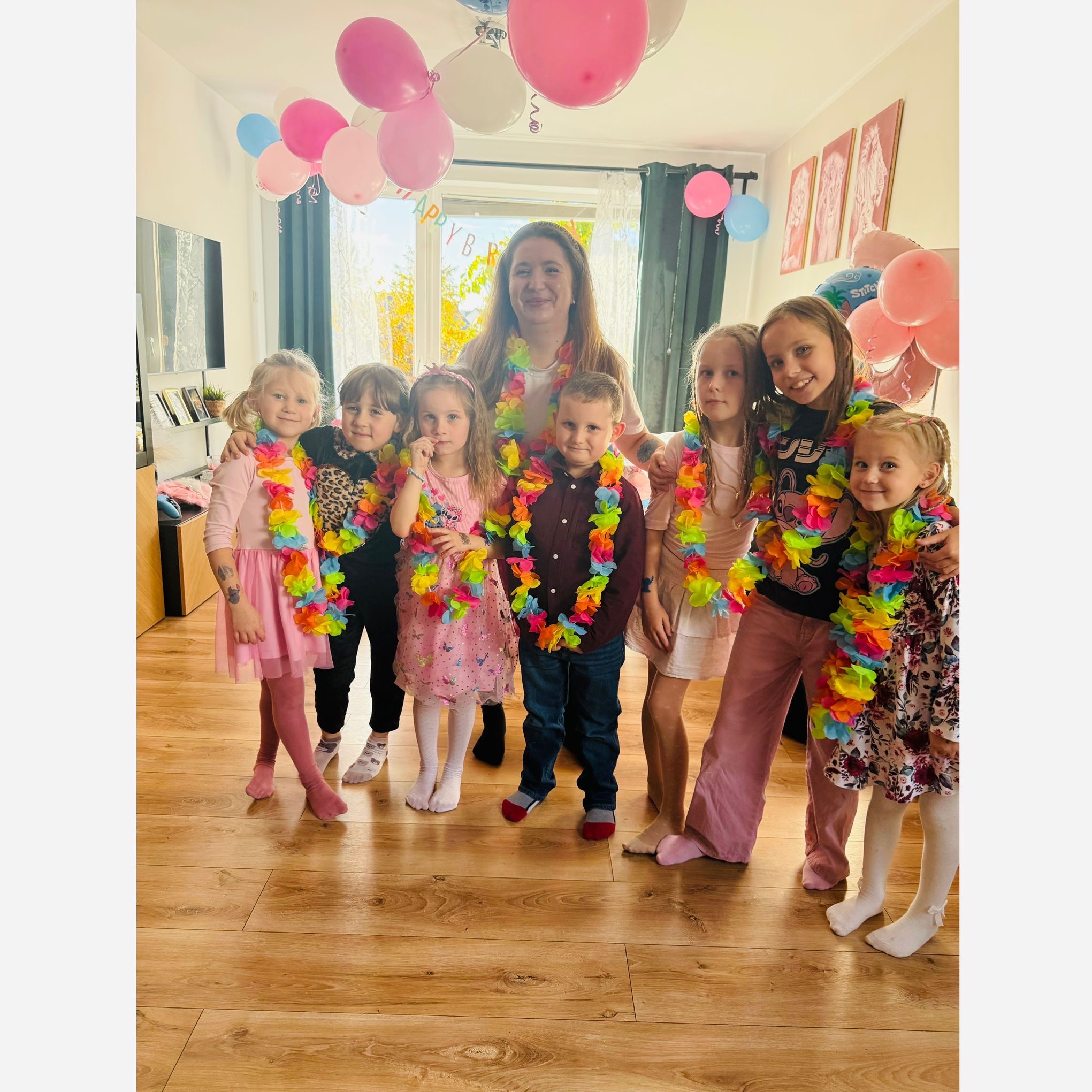 Uśmiechnięta kobieta otoczona grupą dzieci z kolorowymi hawajskimi naszyjnikami na tle pokoju udekorowanego balonami na przyjęcie urodzinowe.